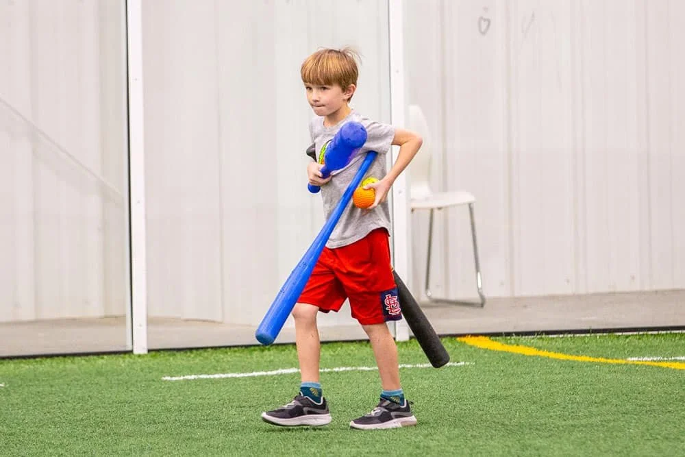 Young boy practicing baseball indoors with a blue bat and helmet, wearing gray t-shirt and red shorts, on artificial grass.