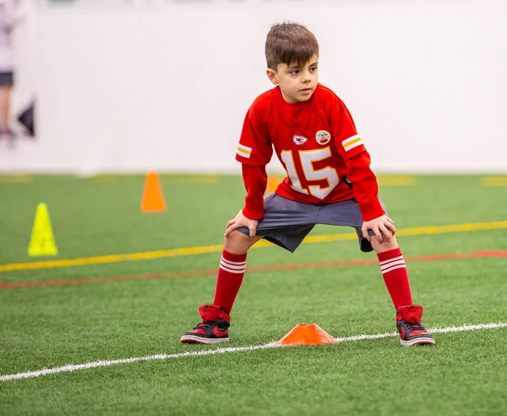 Young boy in a red football uniform practicing agility drills on indoor turf, with orange cones for training.