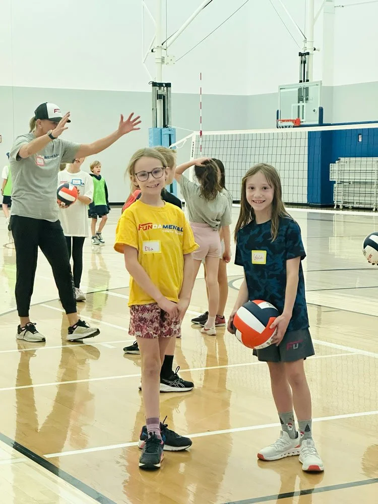 Children and adults playing volleyball in an indoor gymnasium.