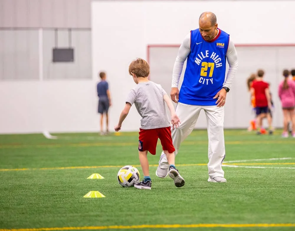 A coach and a young boy practicing soccer on an indoor field, with other children in the background.