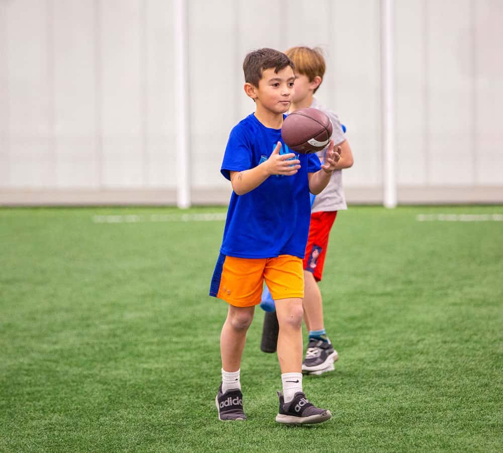 Young boy in a blue t-shirt and orange shorts holding a football indoors on a green turf field, with another child in the background.