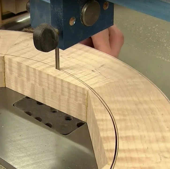 Close-up of a woodworking bandsaw cutting a curved line on a piece of light-colored wood.