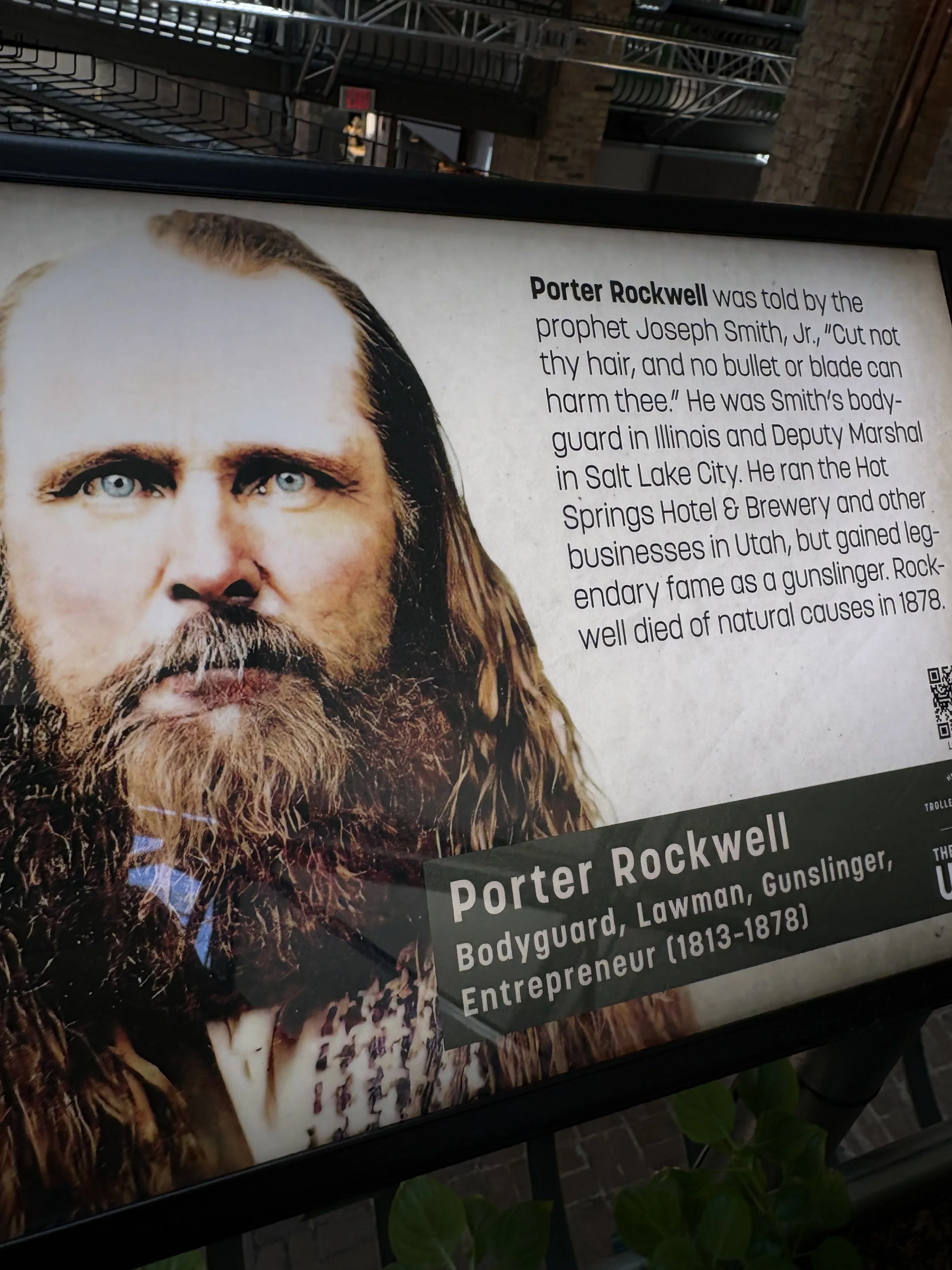 Sign with a portrait of Porter Rockwell, a rugged man with long hair and a beard. The sign includes biographical information about him, noting his role as a bodyguard and lawman in Utah and Illinois, and his reputation as a gunslinger and entrepreneur in the 1800s.