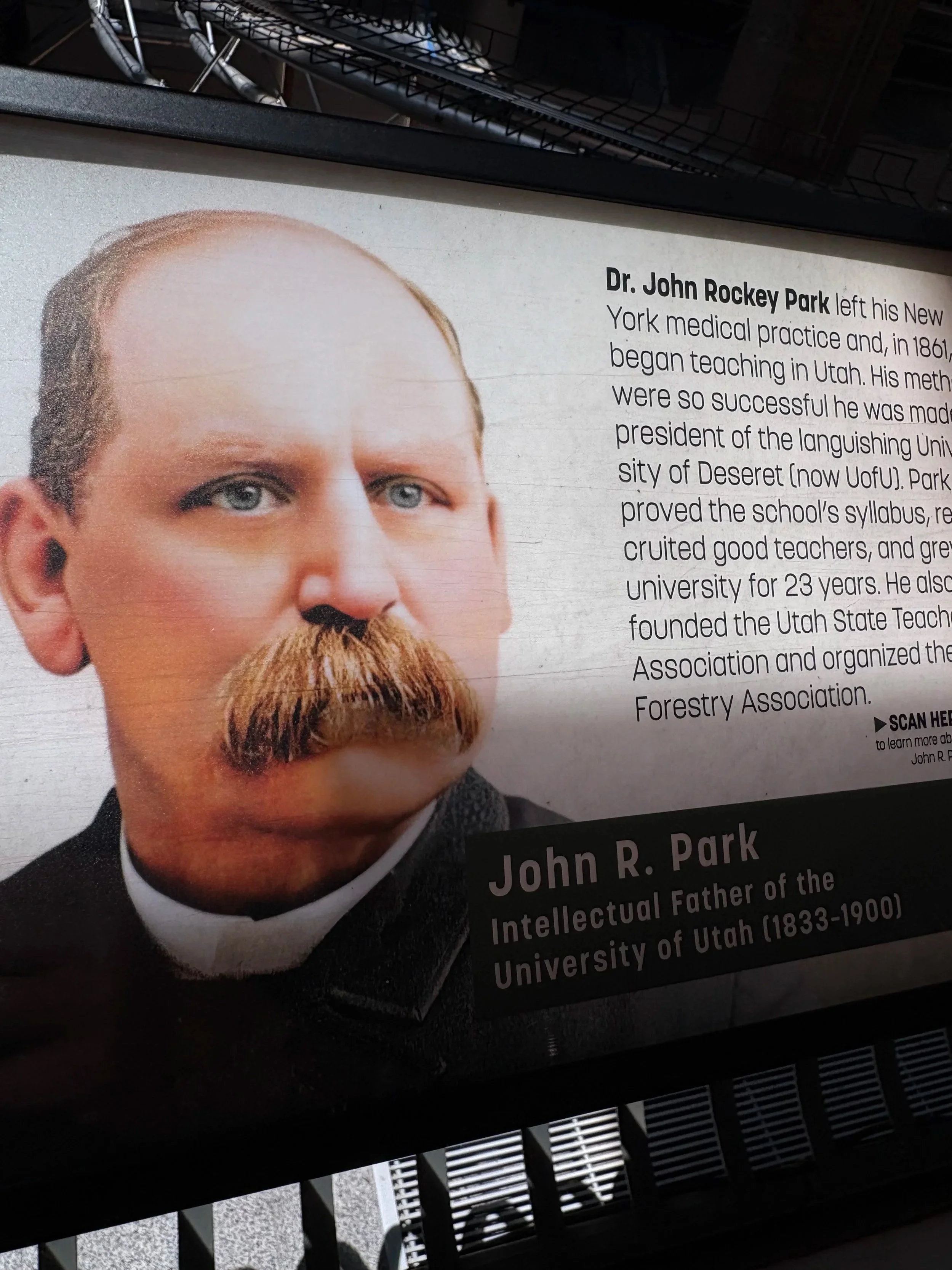 A historical plaque featuring a portrait of Dr. John Rockey Park and text about his contributions, mounted outdoors with shadows from a nearby structure.