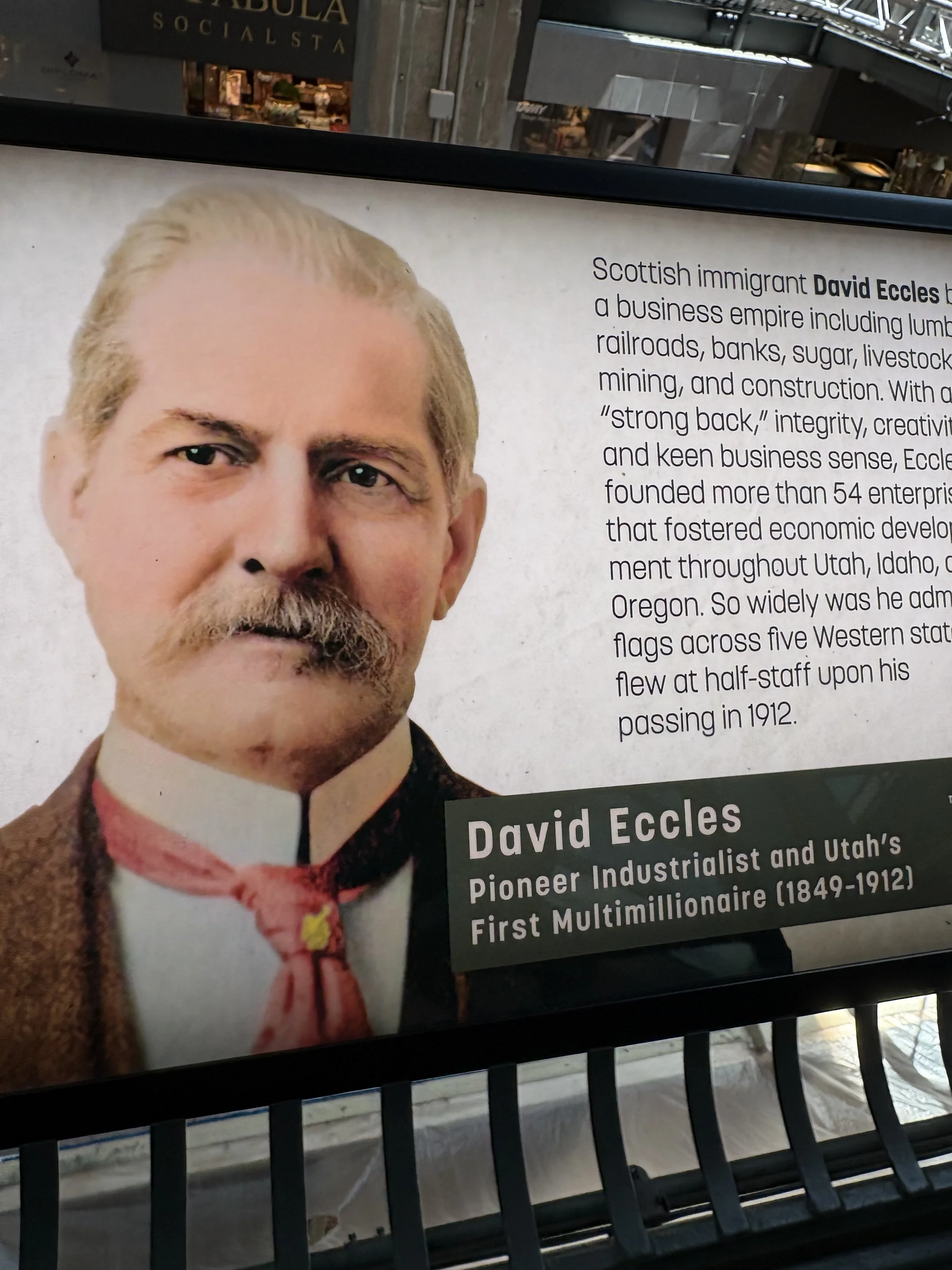 A sign with a portrait of a man with light-colored hair and beard. The sign includes a biography indicating he is David Eccles, an industrialist from Utah who was a pioneer and first multimillionaire. The background shows parts of a mall or shopping center.