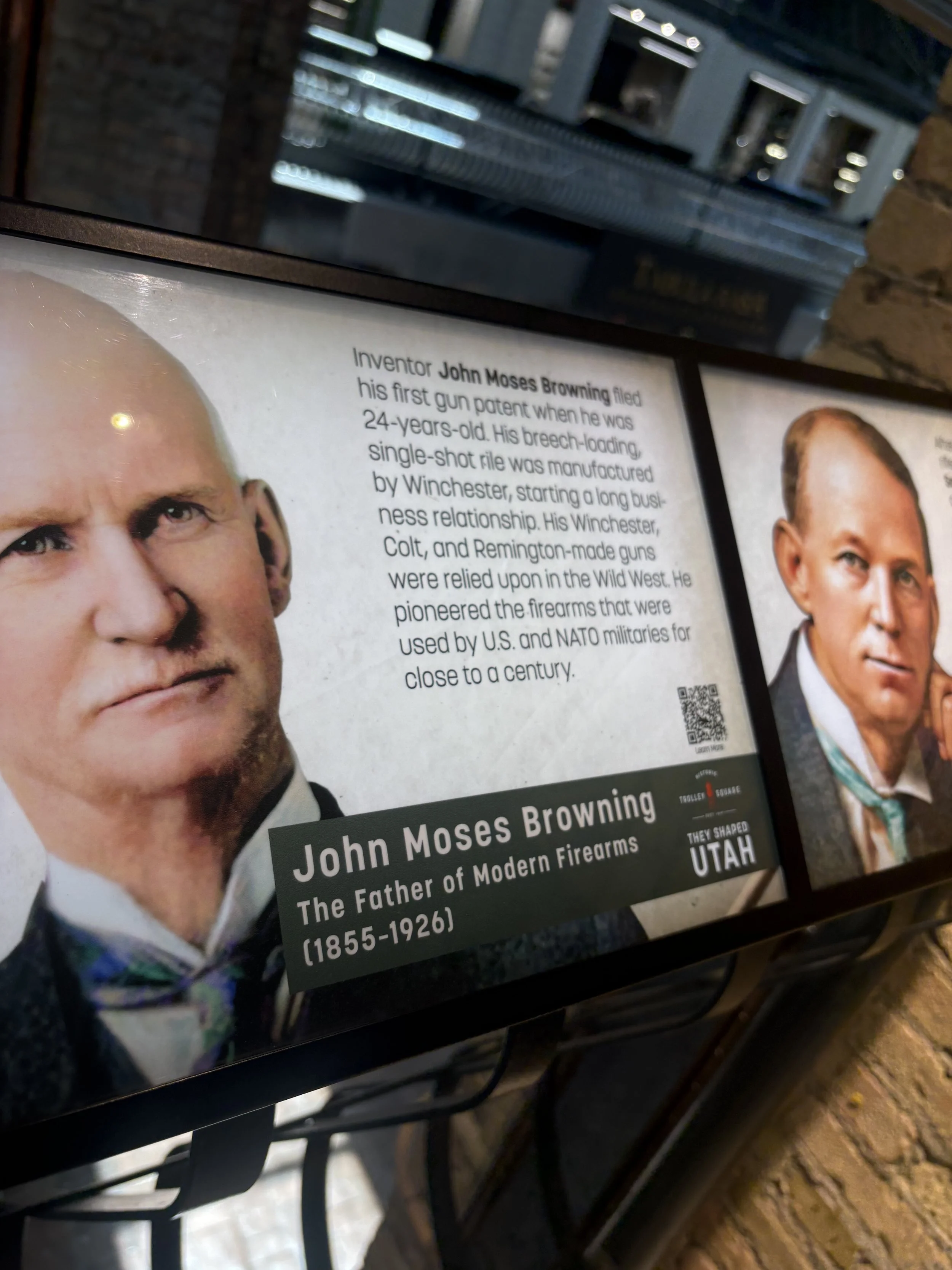 Photograph of informational display about John Moses Browning, featuring his portrait and biography text. The display is mounted on a wall or stand with a brick wall background, and part of a metal grid or structure is visible in the foreground.