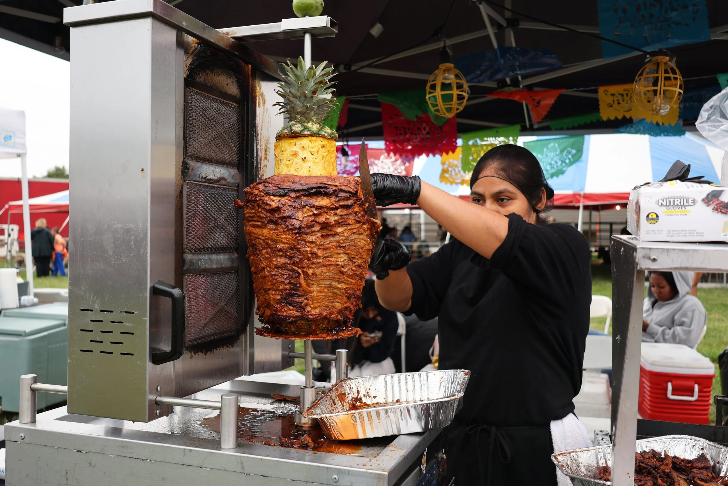 A woman in a black shirt and black gloves preparing a large vertical spit of roasted meat at an outdoor food stand, with a pineapple with a yellow top on top of the spit, under a tent decorated with colorful paper banners.