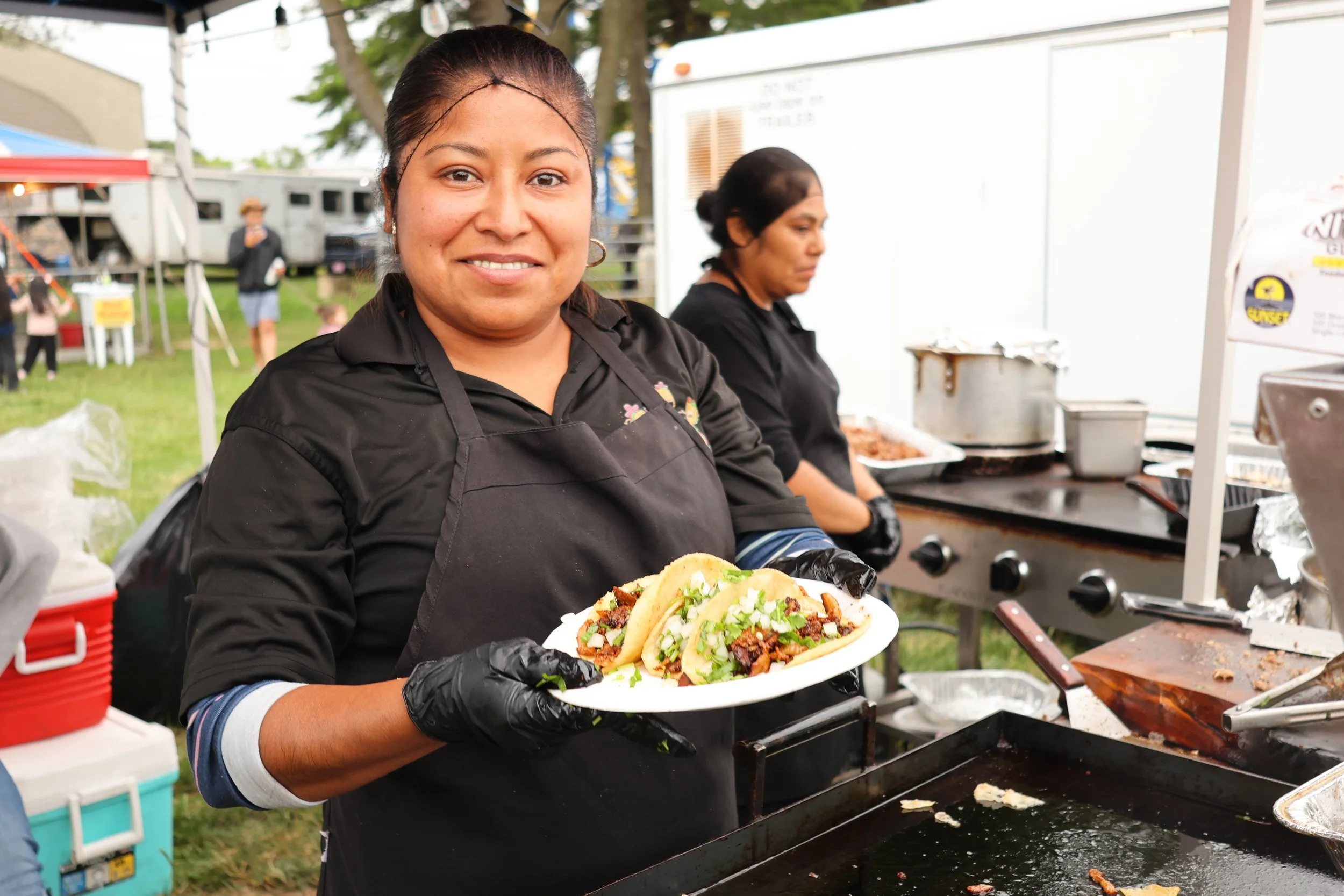 A woman wearing a black apron and black gloves holds a plate of tacos at an outdoor event, with another woman working at the grill in the background.