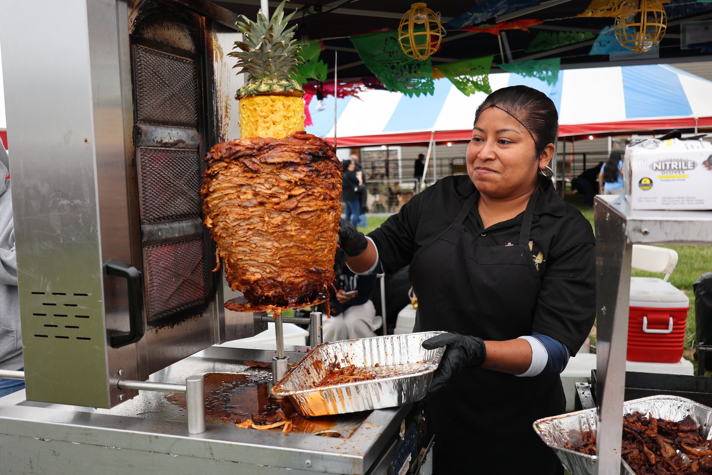 A woman is preparing a large spit of roasted meat at an outdoor event, with colorful decorations and a pineapple on the spit, and other people in the background.