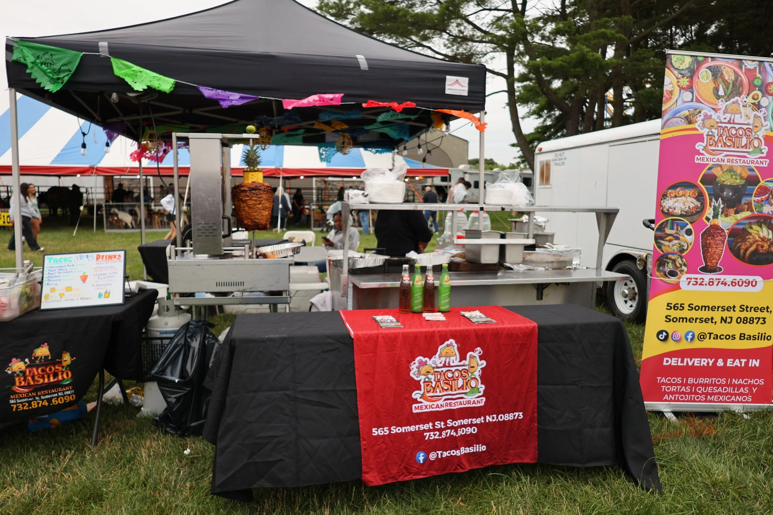 A food stand at an outdoor event featuring a black canopy with colorful papel picado decorations, a banner with the restaurant's name 'Tacos Basilio' and contact information, including address and phone number, and tables displaying menu items like tacos, burritos, and nachos. In the background, there are trees, a red and white tent, and people walking around.