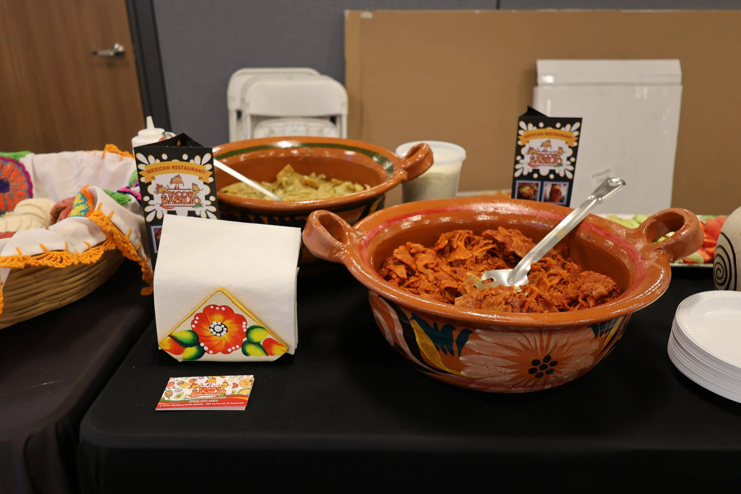 A table with traditional Mexican dishes in colorful pottery, including shredded meat and a creamy dip, along with baskets of tortillas and a small flag with flowers and Mexican restaurant branding.