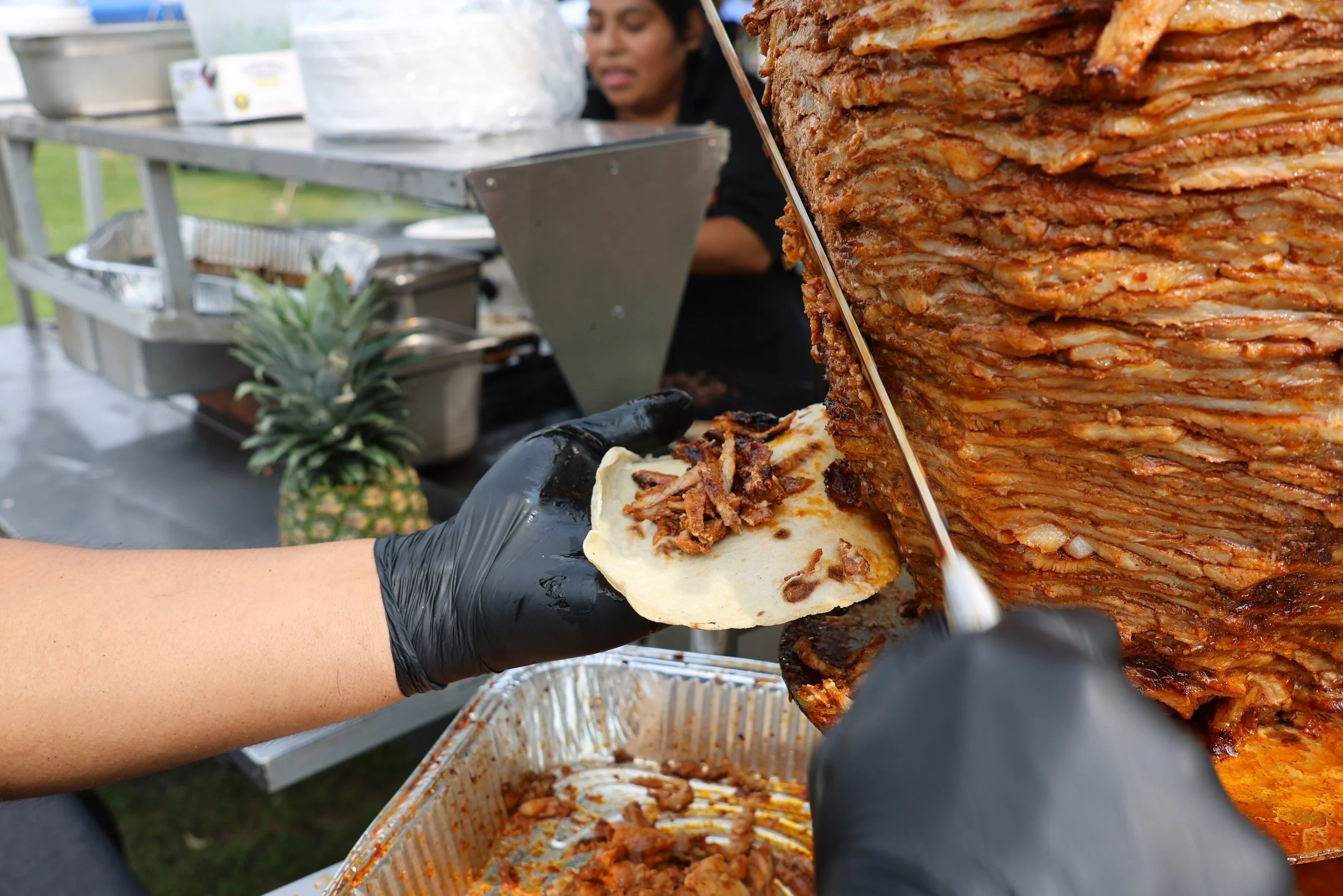 Person slicing a large stack of cooked pulled pork, with a pineapple visible in the foreground and a person in the background near a buffet table.