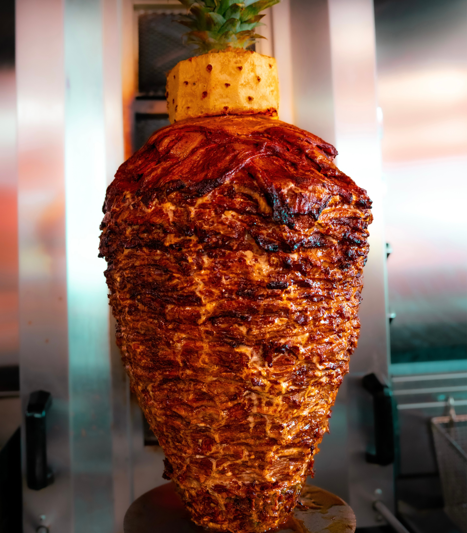Vertical close-up of a large piece of rotisserie spit roast with a crispy, browned exterior, topped with a pineapple on a skewer, in a commercial kitchen or restaurant setting.