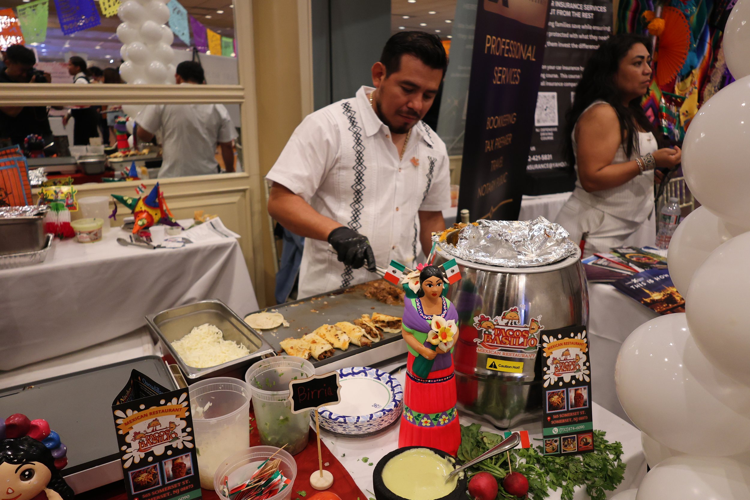 A man in a white embroidered shirt serving food at a Mexican restaurant booth, with various dishes, decorations, and promotional signs in the background.