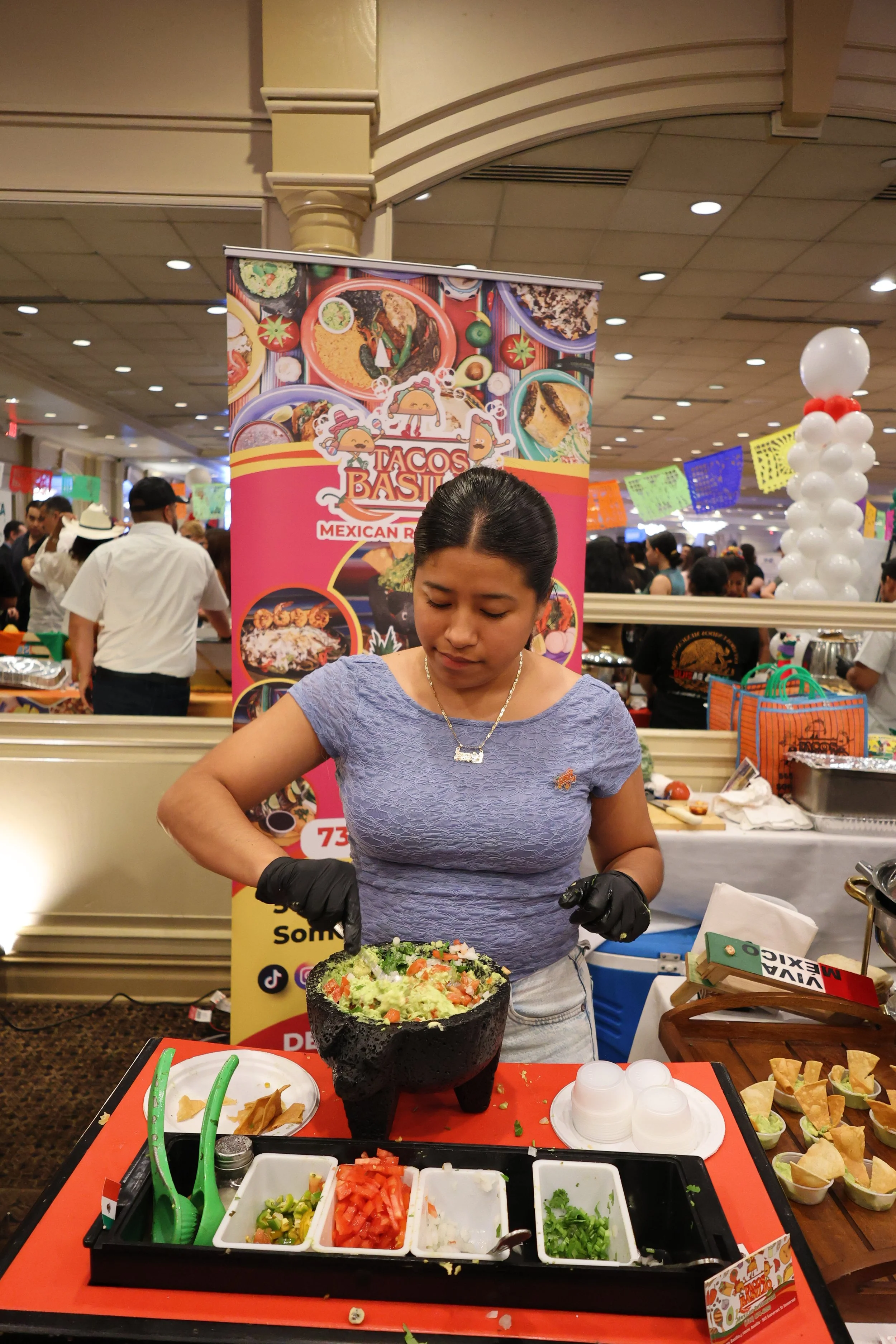 A woman preparing sushi at a food booth during a celebration or festival. She is wearing black gloves and standing behind a table with various ingredients and utensils. Behind her, a bright pink banner with restaurant branding and colorful images of 