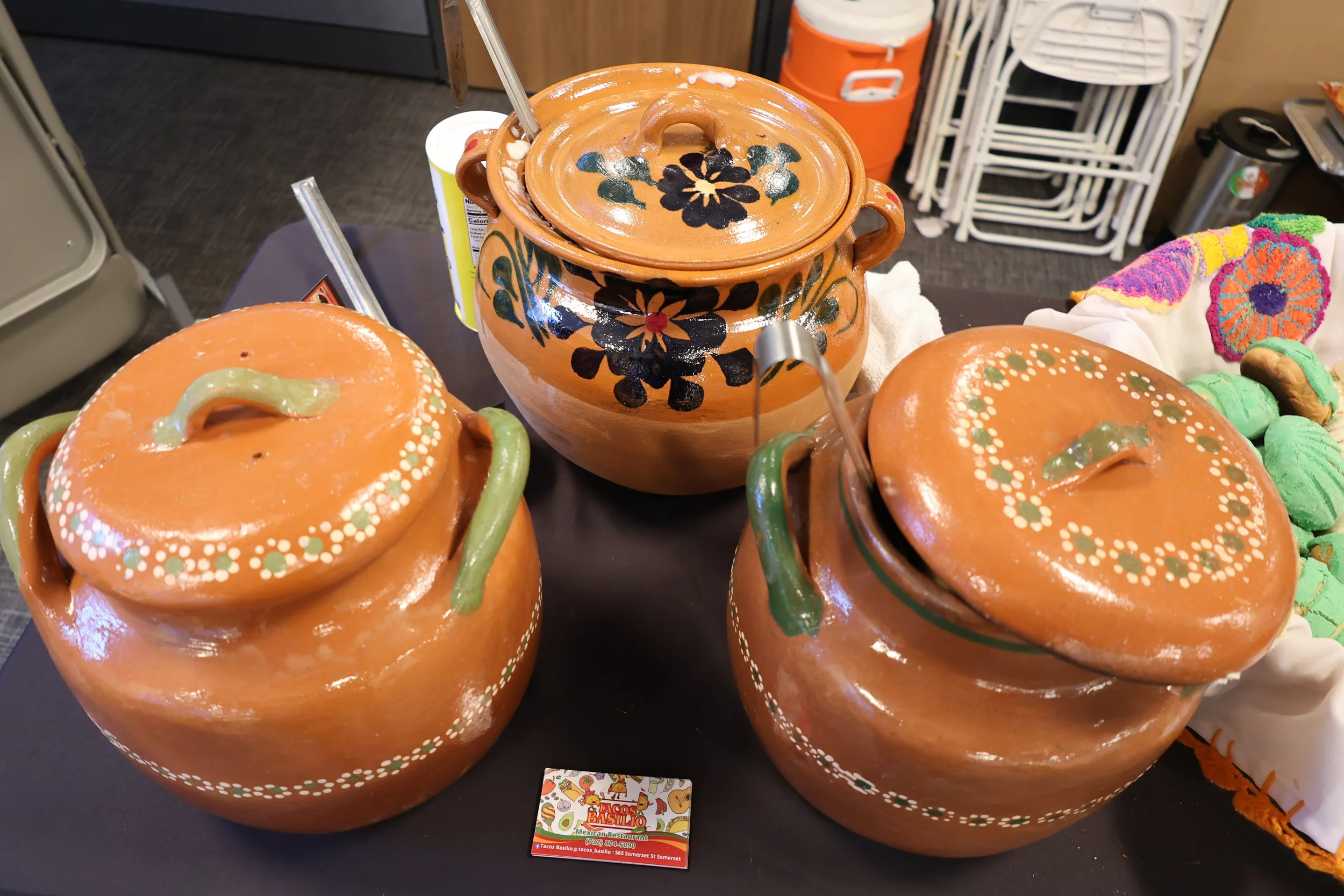 Terracotta Mexican-style soup tureens with painted floral designs, displayed on a black table at a market or shop. In the background, there are stacked white chairs, an orange cooler, a stainless steel trash can, and a colorful embroidered textile.