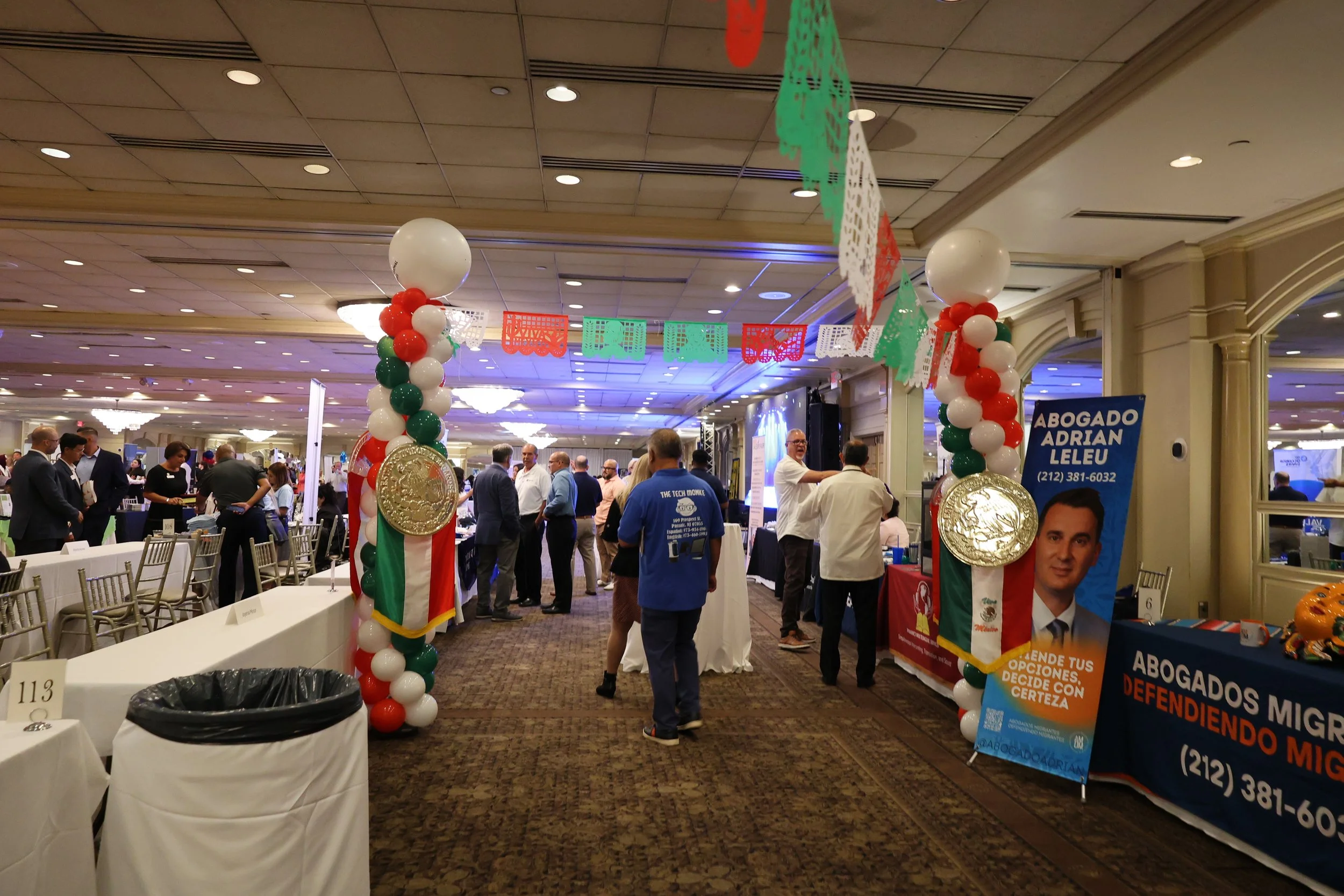 An indoor event decorated with balloons and papel picado banners in green, white, red, and orange, with people engaging at booths and tables in a large, well-lit banquet hall.