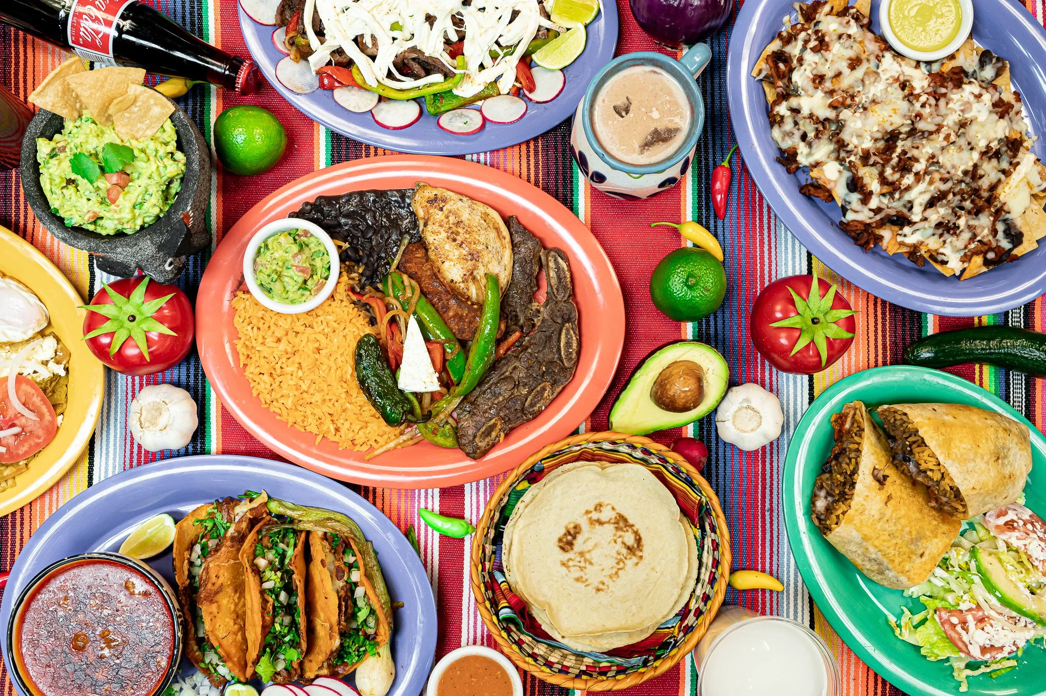 A colorful array of Mexican dishes including tacos, rice, beans, guacamole, nachos, and various side dishes, all arranged on a vibrant striped tablecloth.