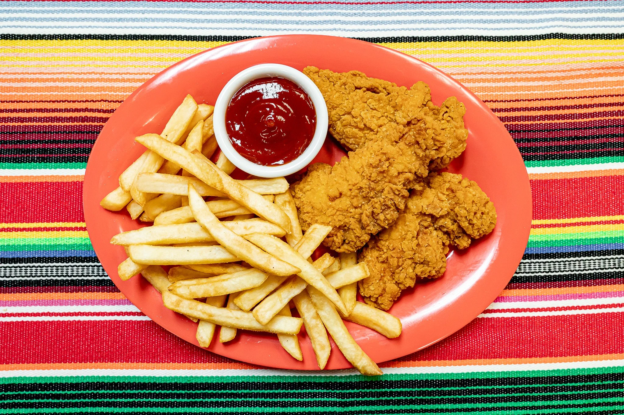 Plate with fried chicken, French fries, and ketchup on a colorful striped tablecloth.