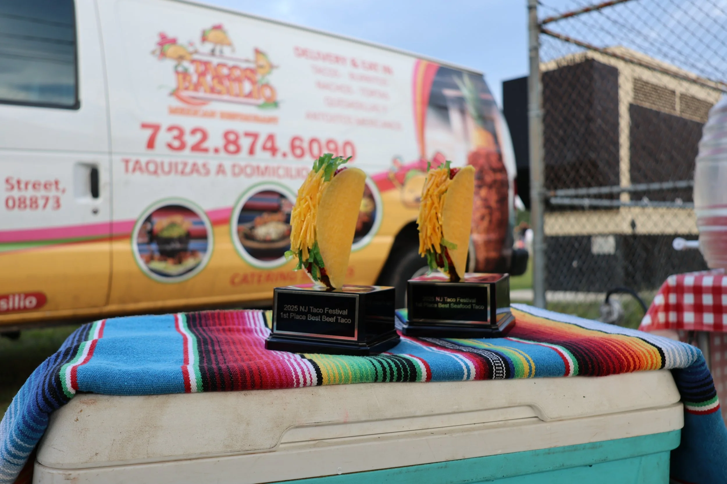 Two taco-shaped trophies on a colorful, striped tablecloth at a taco festival event, with a food truck in the background advertising tacos and a prize for best seafood taco.