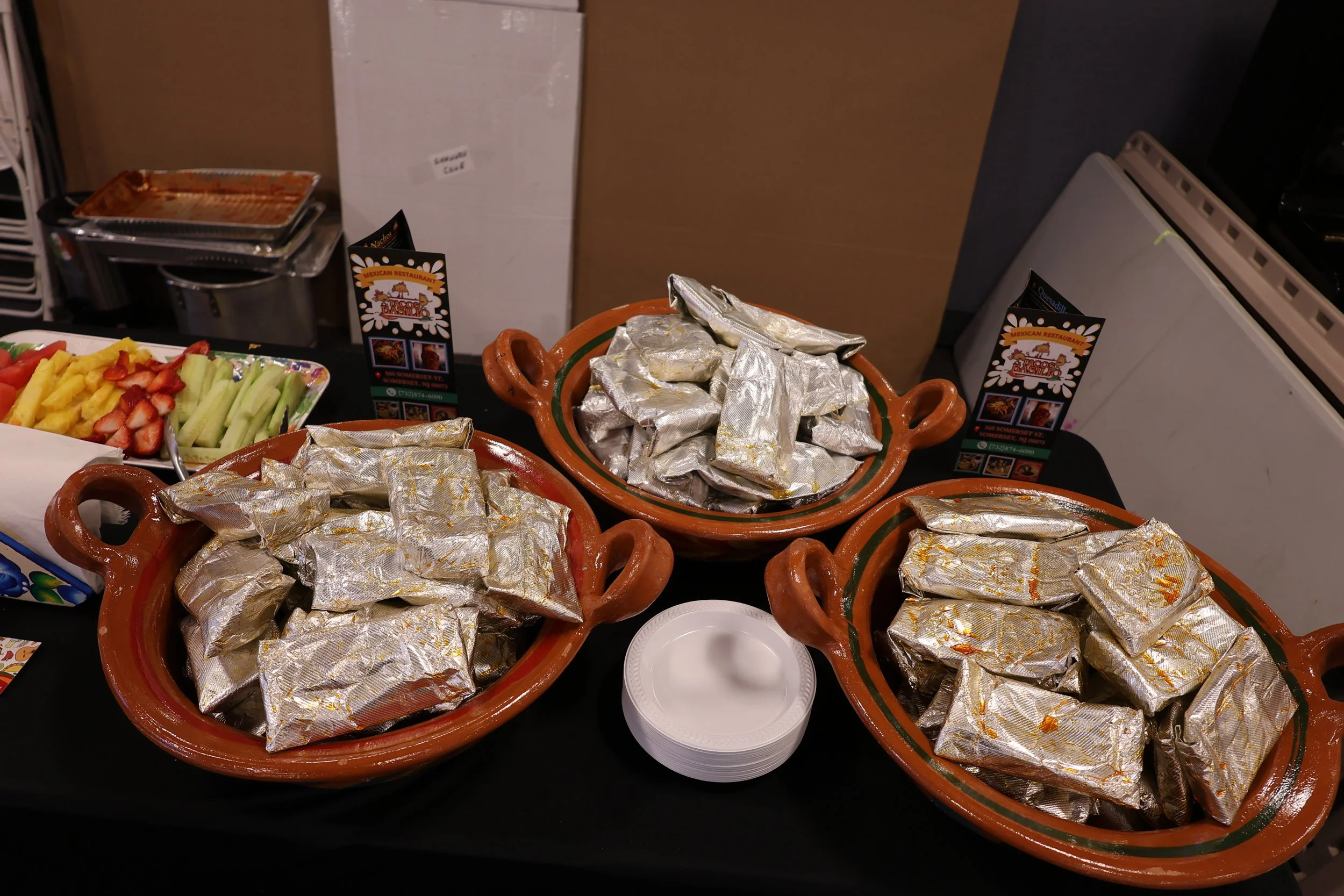 Multiple terracotta pots filled with wrapped aluminum foil-covered dishes, alongside a tray of sliced strawberries and cantaloupe, with a black table surface and business cards for a Mexican restaurant.