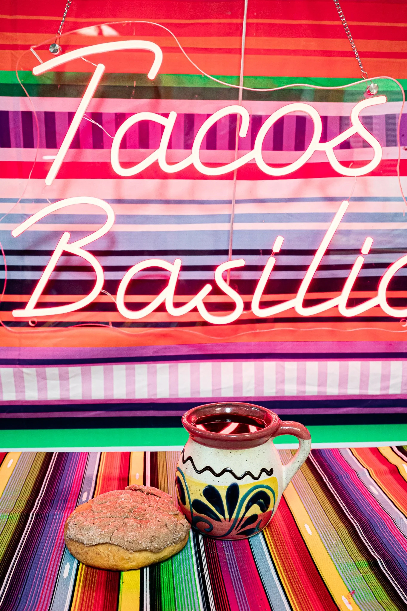 Neon sign that reads "Tacos Basiles" against a colorful striped background with a ceramic mug and a cookie on a multicolored tablecloth.