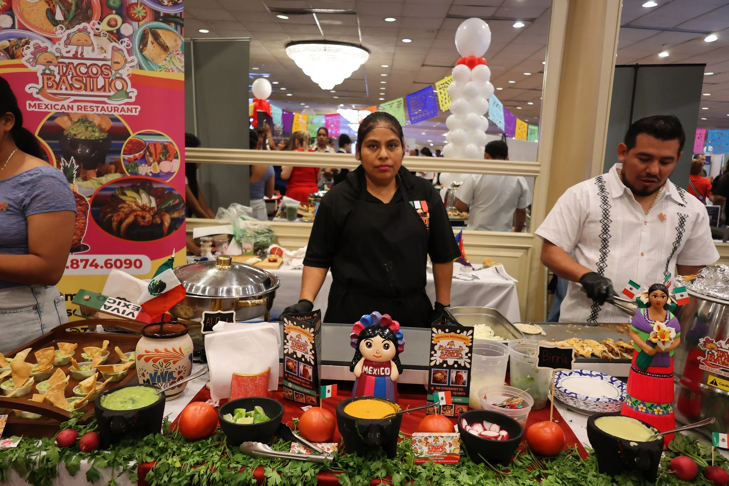 A food stall decorated in Mexican theme with various bowls of dips, vegetables, and nachos, and colorful figurines of Mexican characters, located at an event with people in the background and festive banners hanging from the ceiling.