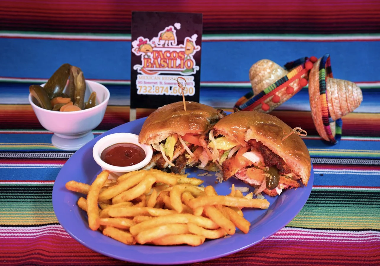 Plate with a cheeseburger, French fries, and a side of ketchup, a bowl of pickles, a small sign for a Mexican restaurant, and colorful sombreros, all on a bright striped tablecloth.