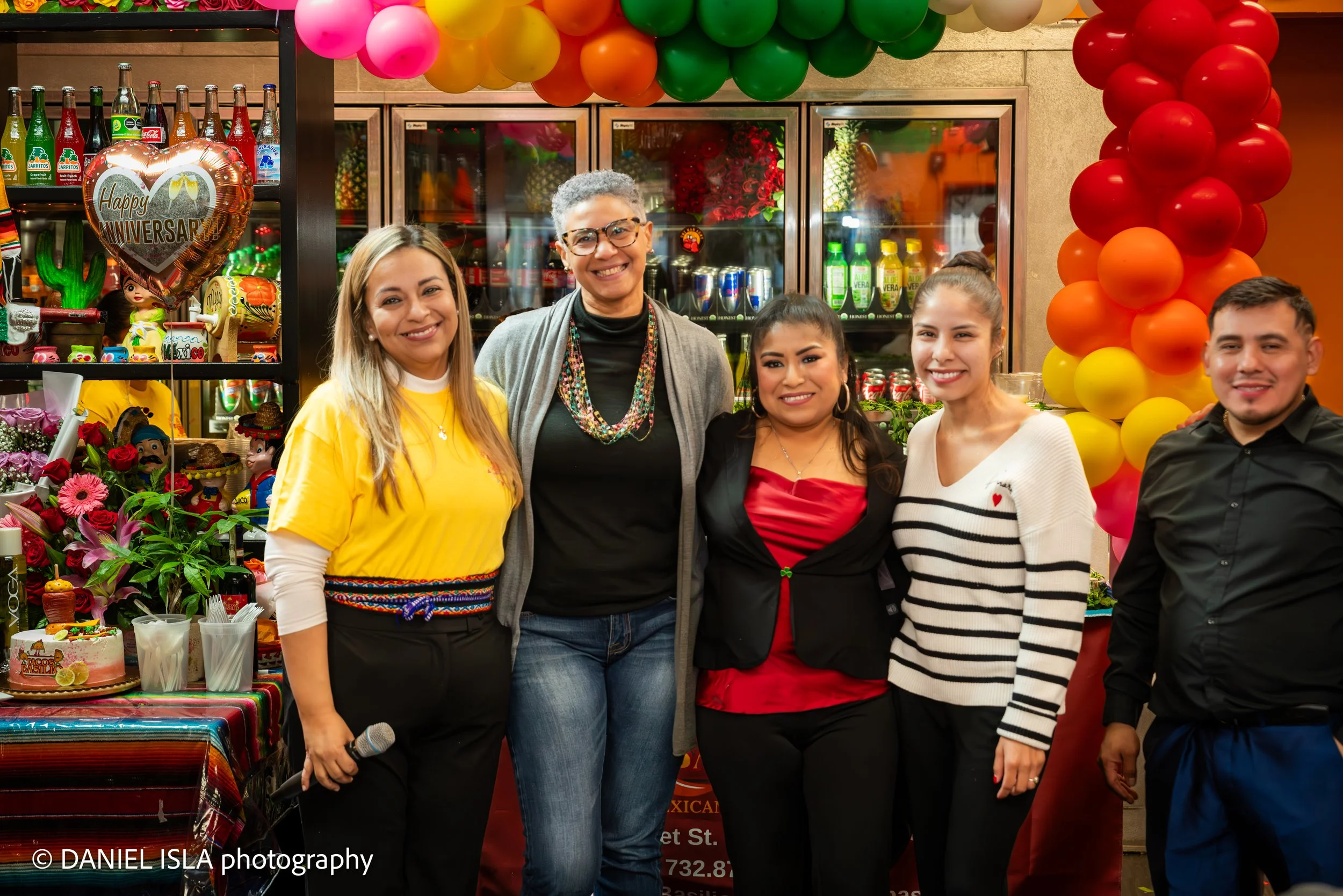 Four women and one man are smiling at a celebration with colorful balloons and decorations, next to a table with flowers and a cake.