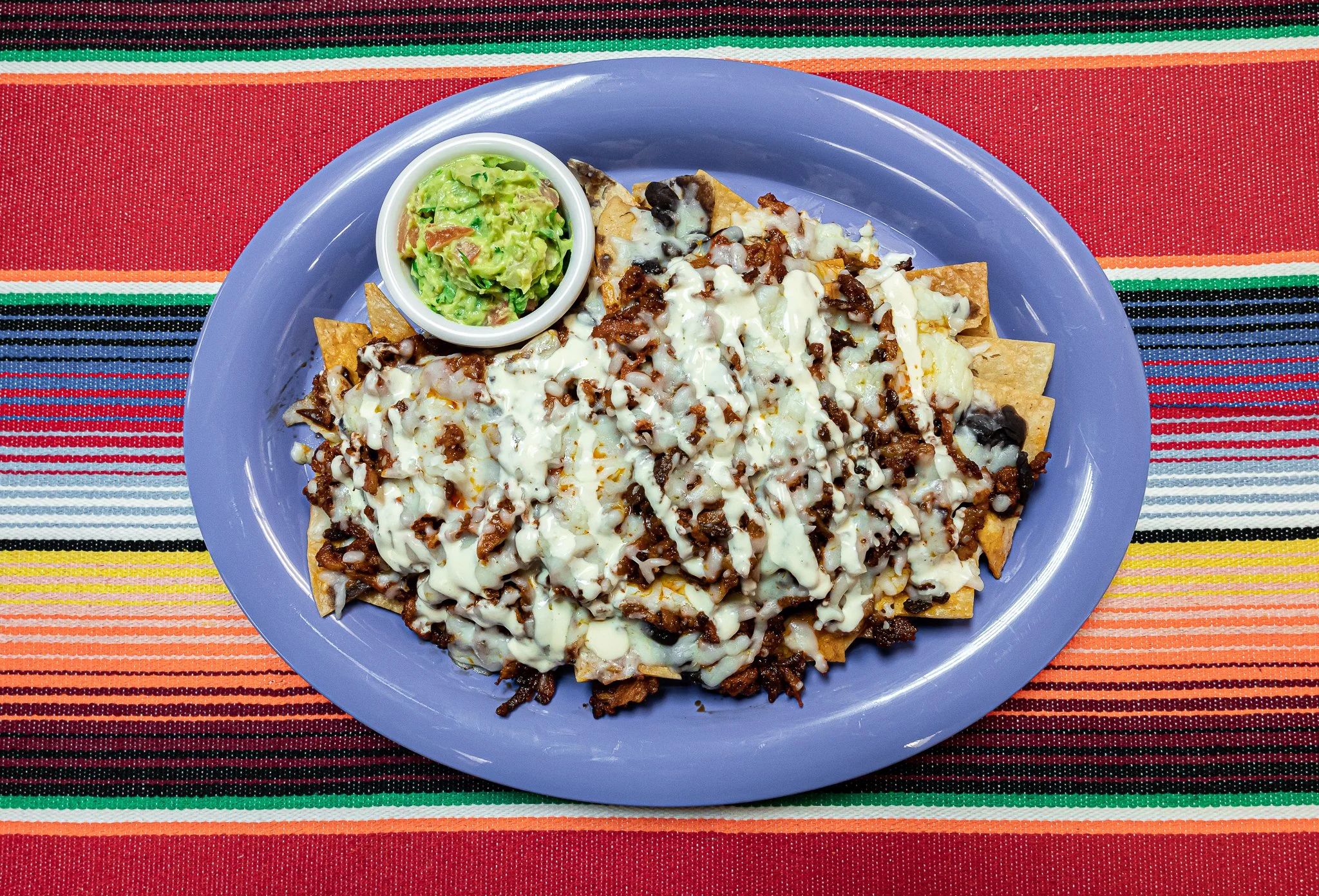 Plate of nachos topped with melted cheese and seasoned meat, served with a side of guacamole in a small white bowl, on a colorful striped tablecloth.