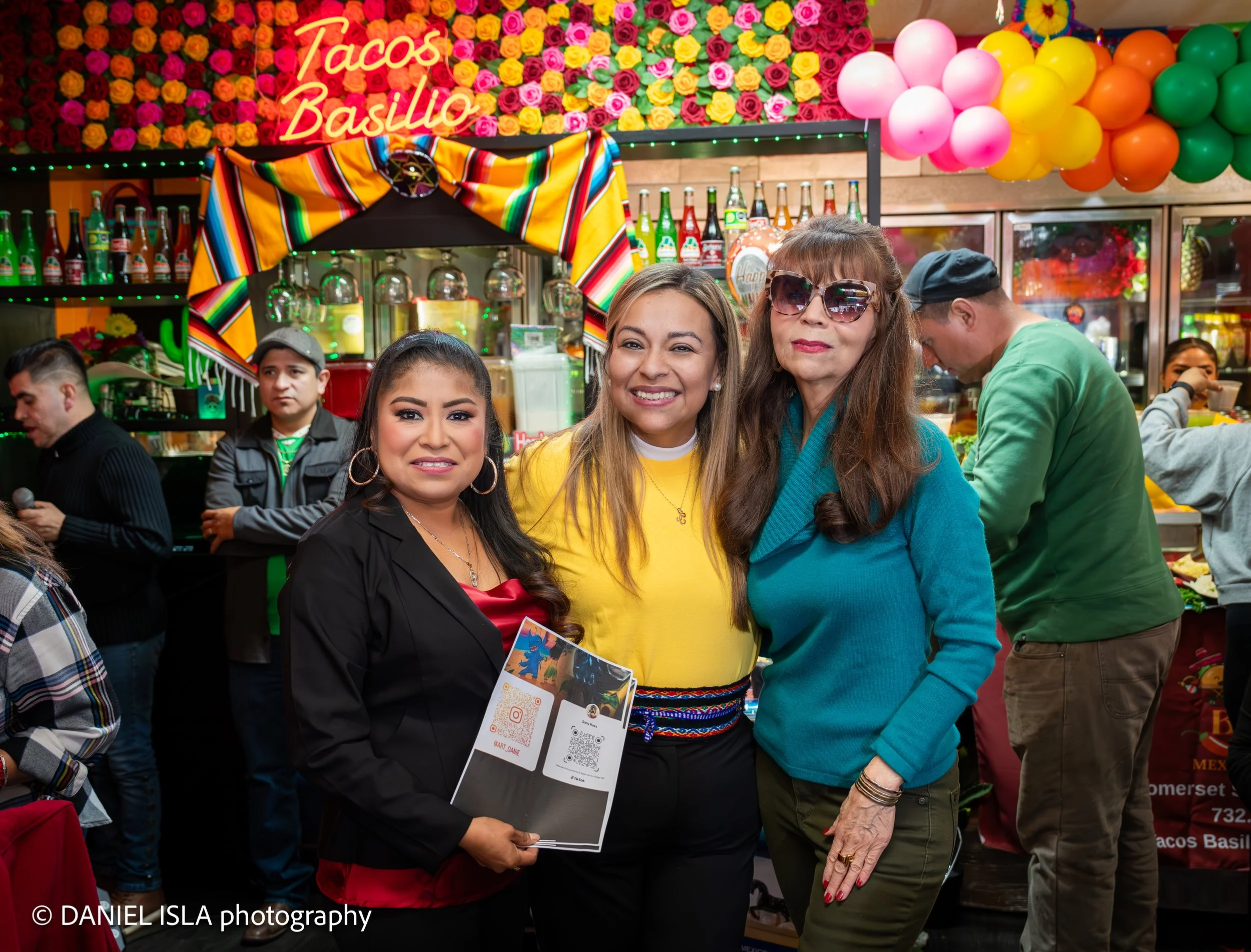 Two women smiling and posing for a photo at a market stall decorated with colorful balloons and flowers, with bottles of Mexican soda behind them and a sign that reads 'Tacos Basilio' in neon letters.