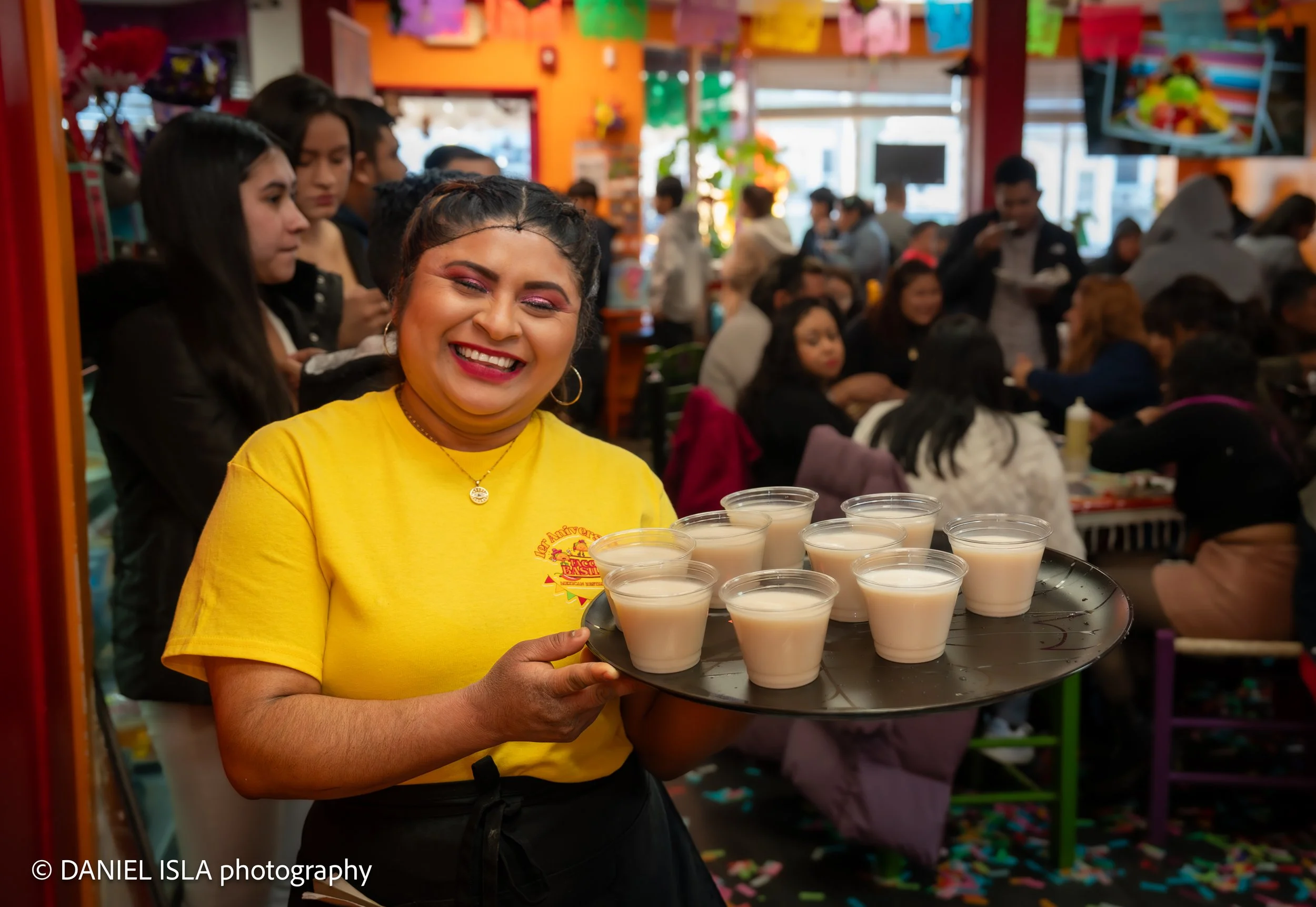 A woman in a yellow shirt holding a tray of white cups filled with a light-colored beverage, with colorful decorations and people in the background