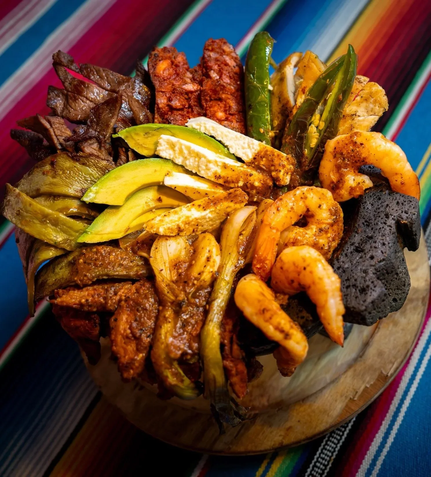 A traditional Mexican dish called 'torta' with various fillings including sliced avocado, shrimp, grilled meats, green chilies, and cheese on a wooden board, with vibrant Mexican textile in the background.