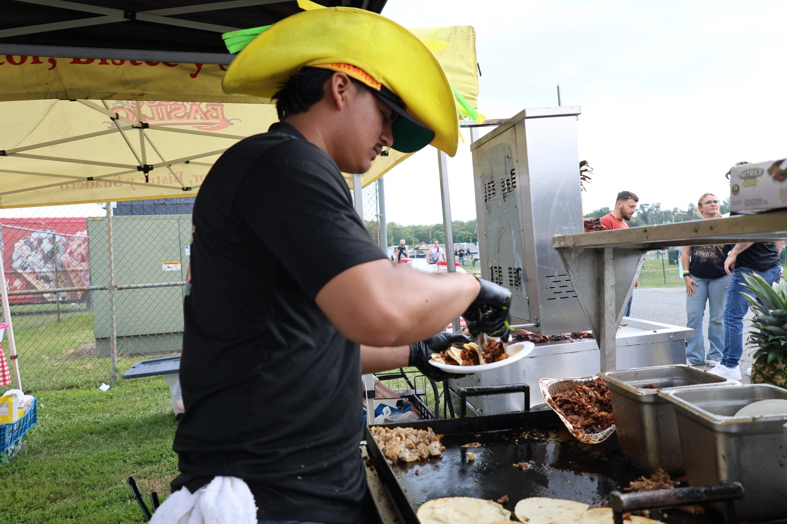 Person serving food at an outdoor barbecue stand, wearing a large banana-shaped hat.