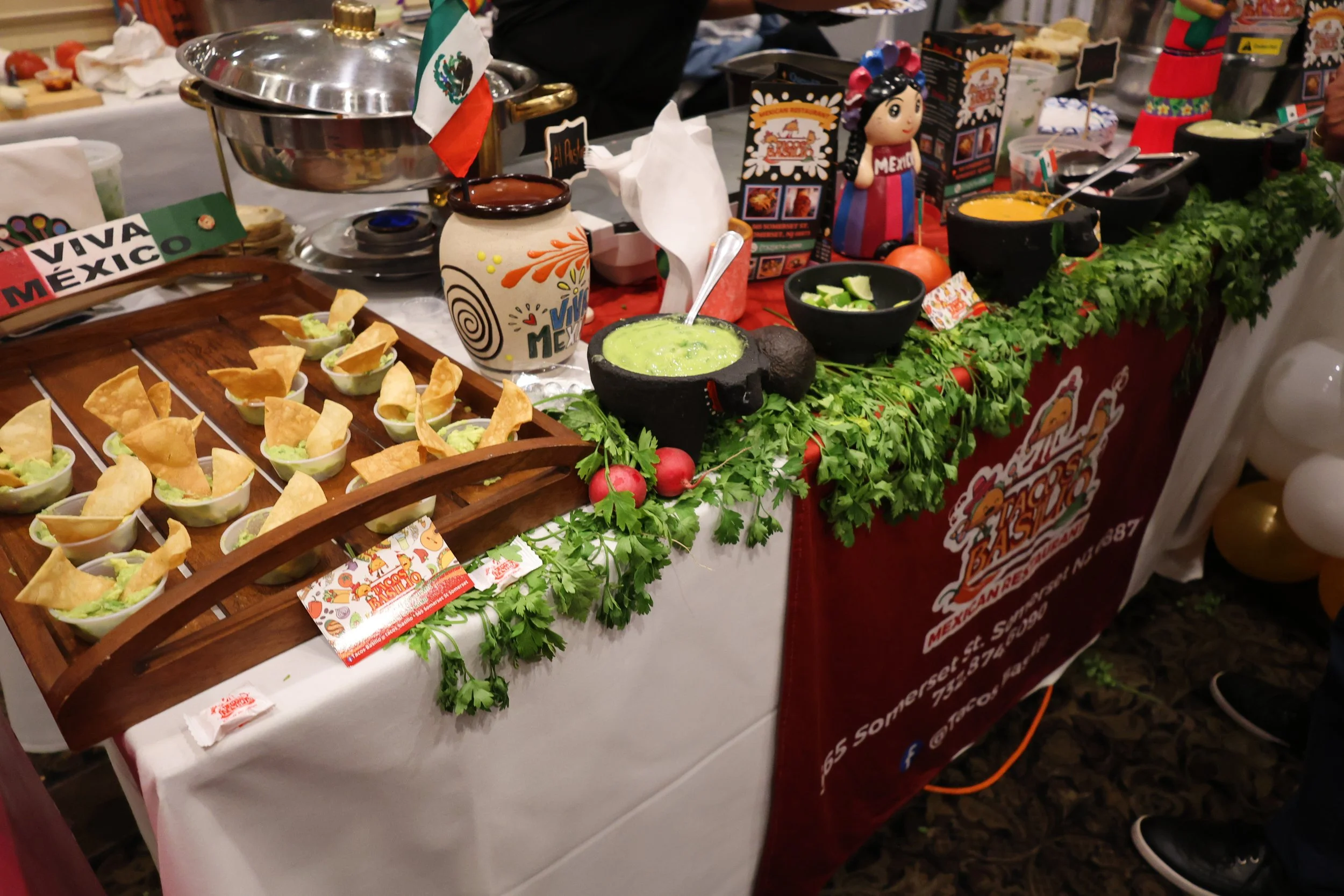 A table decorated with Mexican themed items including small bowls of guacamole with chips, a decorative mug with the words 'Viva Mexico,' salsa, and fresh cilantro. There are also colorful figures and signs promoting Mexican cuisine, indicating a fes