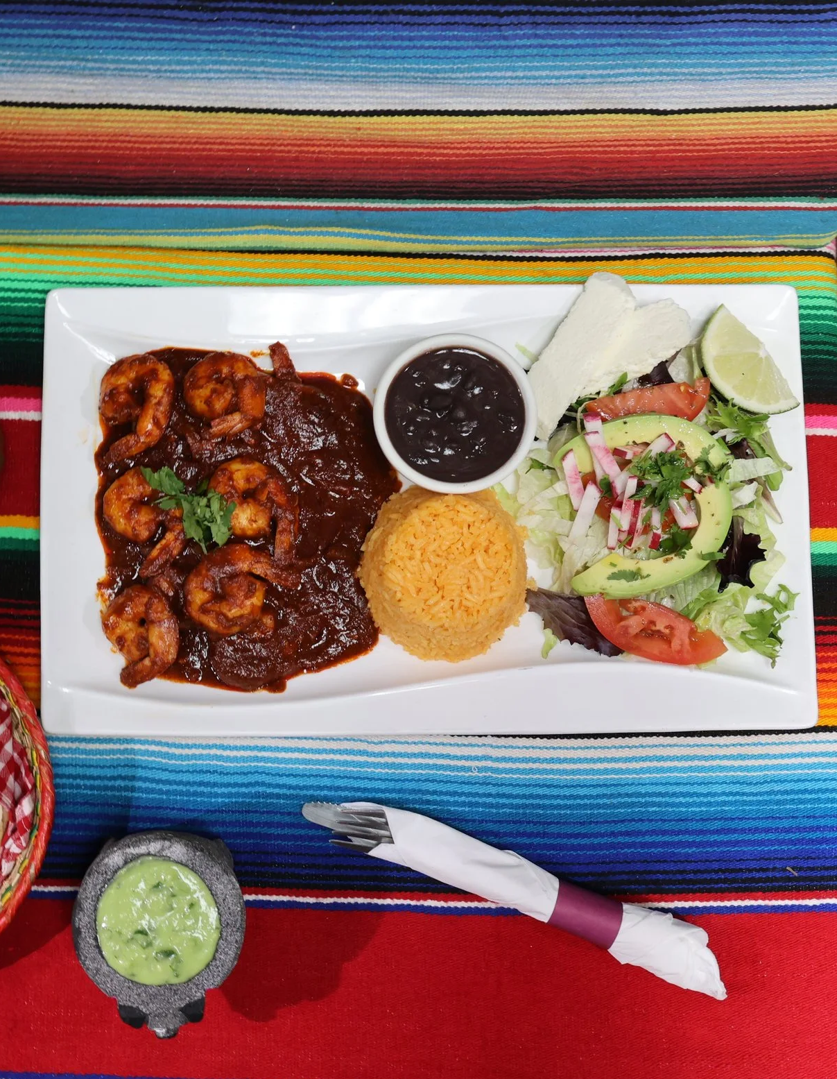 A rectangular white plate with Mexican food including shrimp in dark sauce, Mexican rice, black beans, and a side salad with tomatoes, avocado, and lettuce. Accompanying the plate are lime wedges and a small dish of green sauce, placed on a colorful striped tablecloth.
