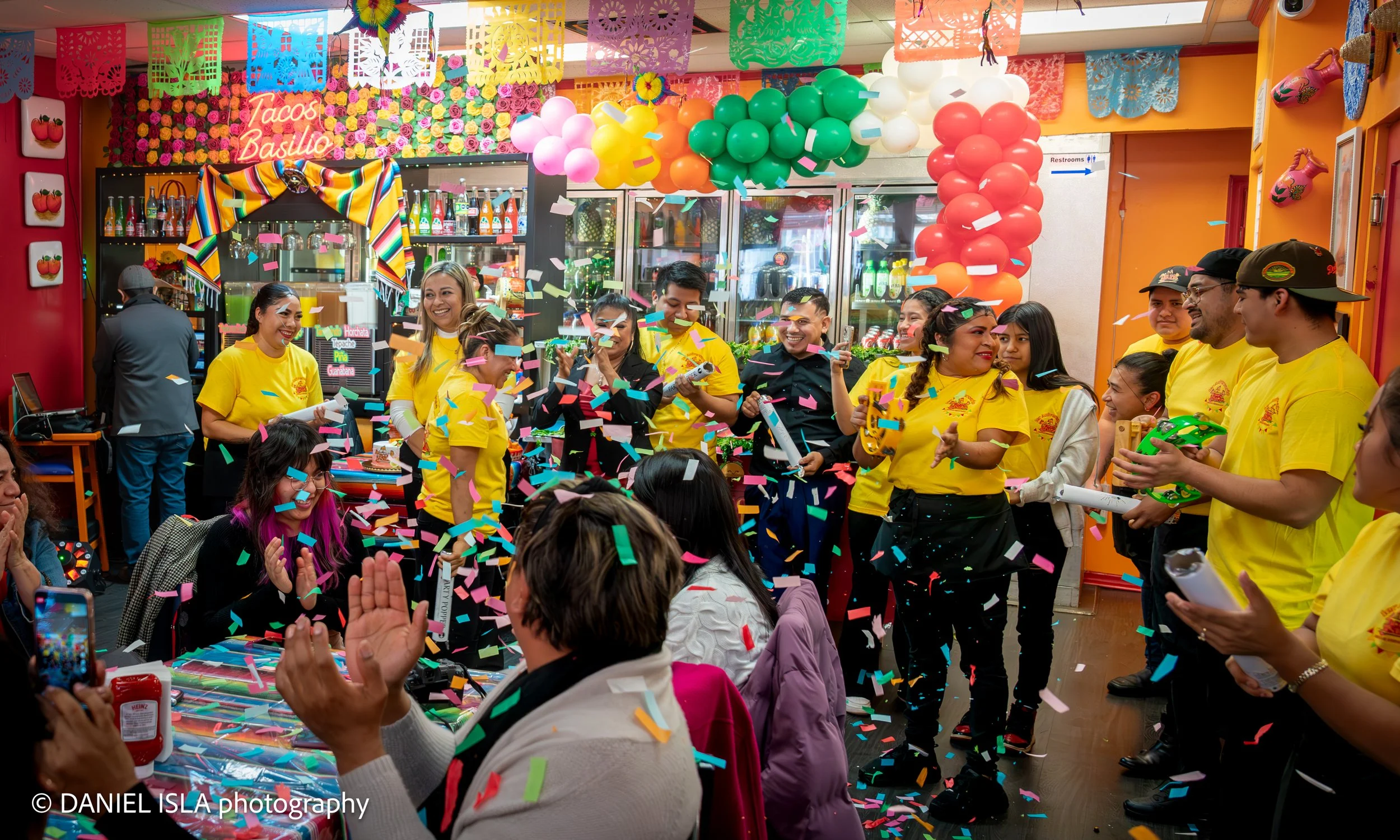 Celebration with colorful balloons, confetti, and smiling people in a lively restaurant or cafe, decorated with vibrant paper banners and floral wall art, with a sign that reads 'Tacos Basilio'.