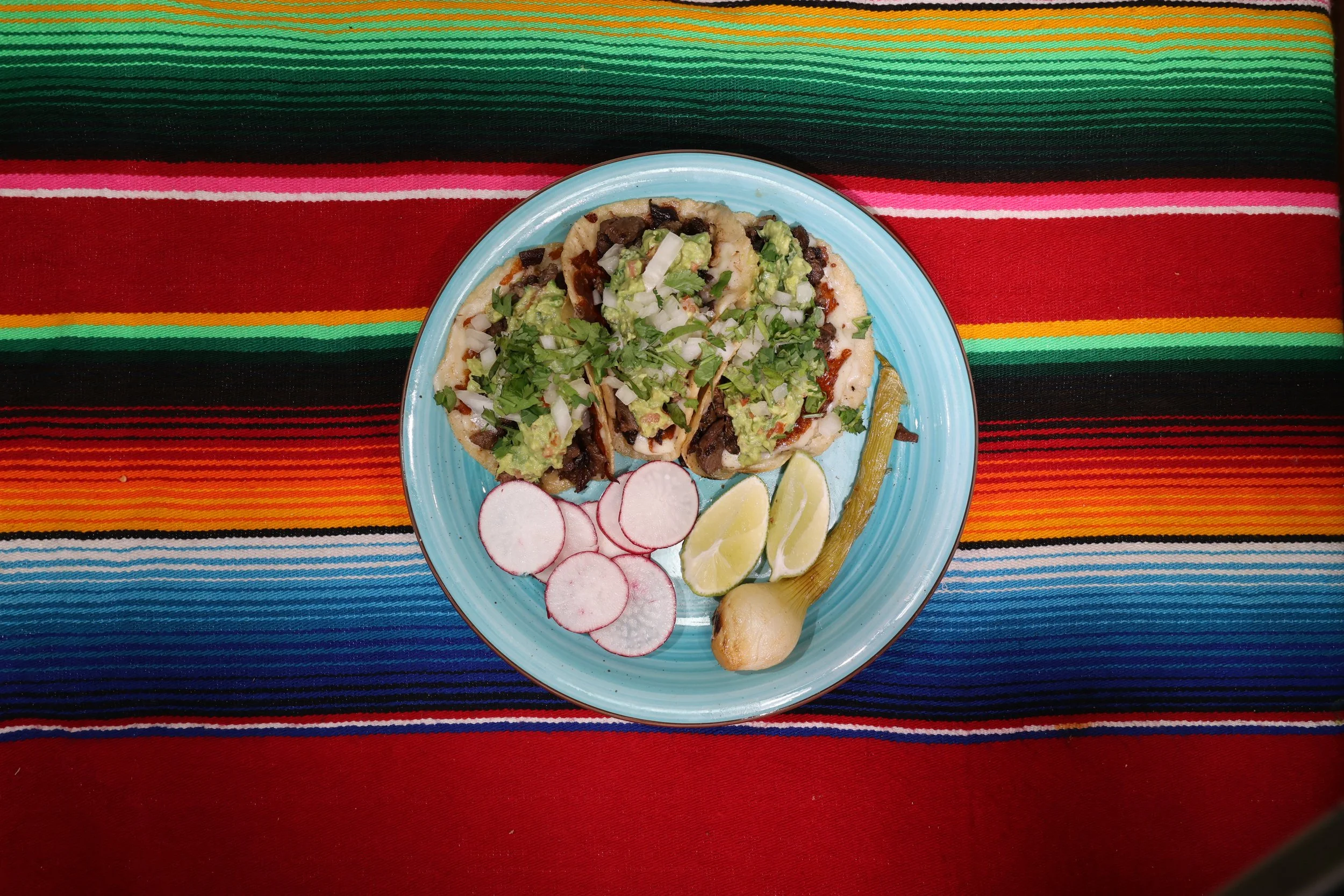 Plate of tacos garnished with chopped onions, cilantro, avocado slices, radish slices, lemon wedges, and a grilled jalapeno pepper, on a vibrant striped textile background.