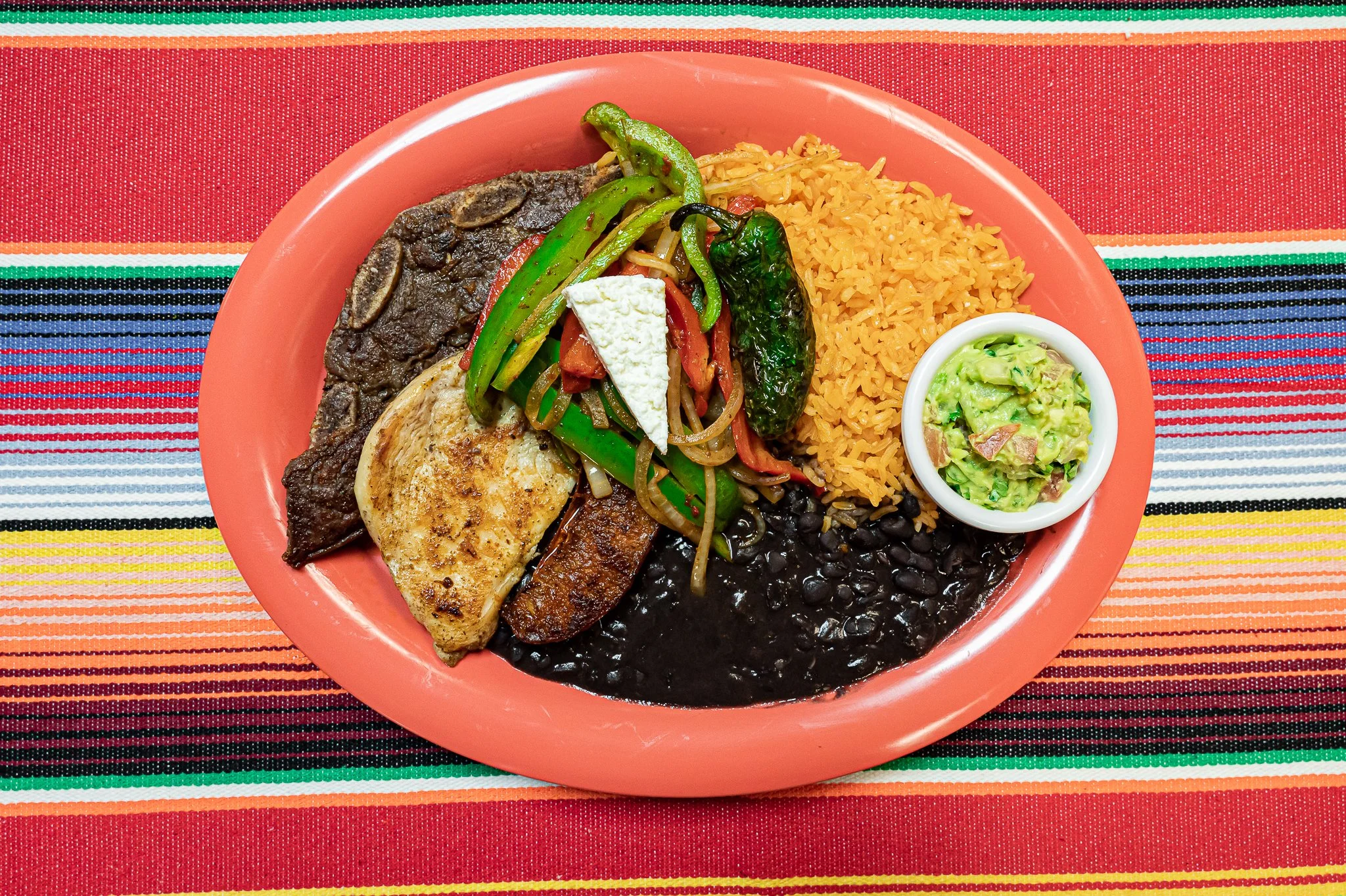 A plate of Mexican food with rice, black beans, grilled chicken, beef steak, grilled peppers, tomato slices, with guacamole on the side, all on a colorful striped tablecloth.
