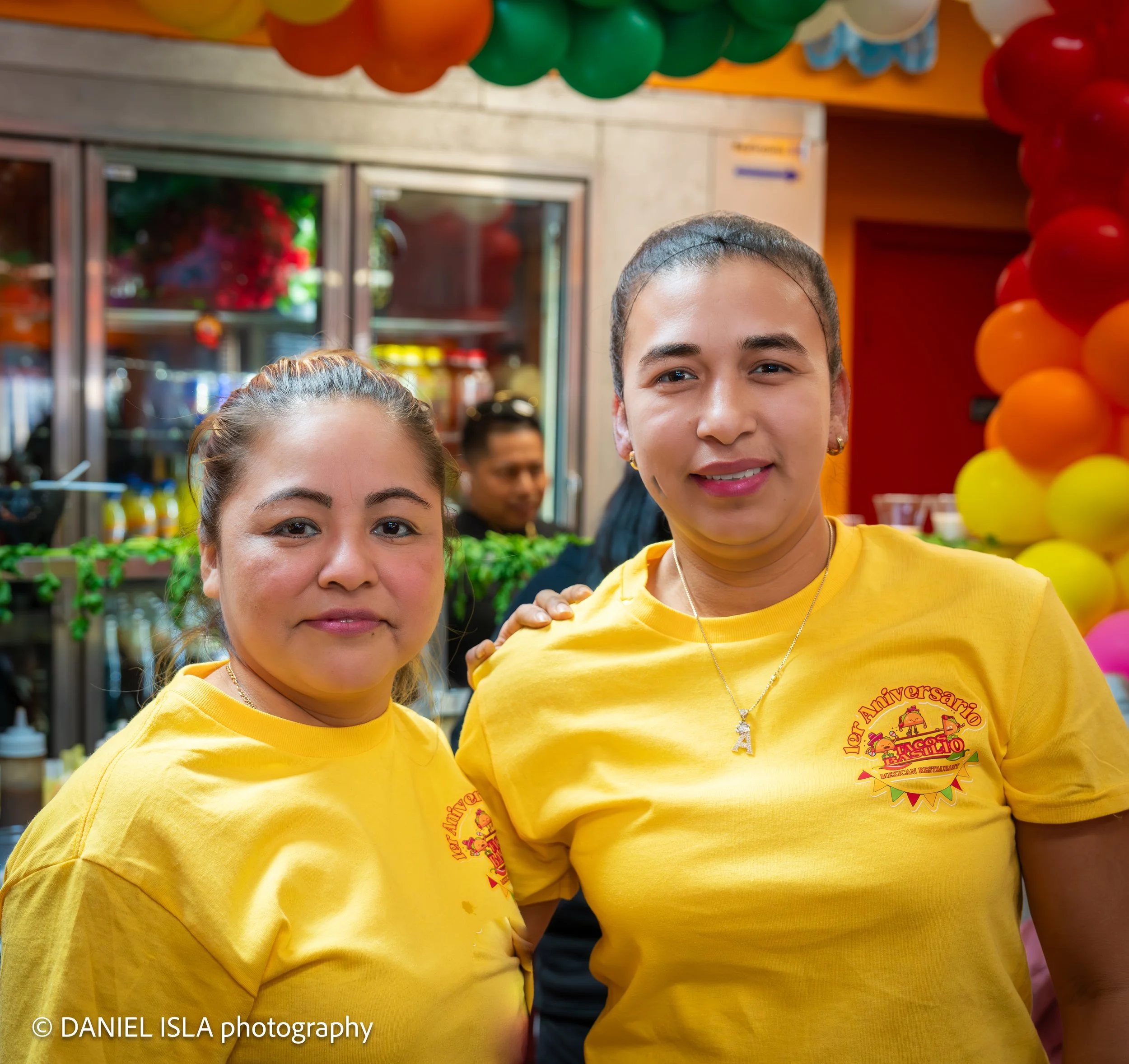 Close-up of two smiling young women wearing yellow shirts at a colorful celebration with balloons in the background.