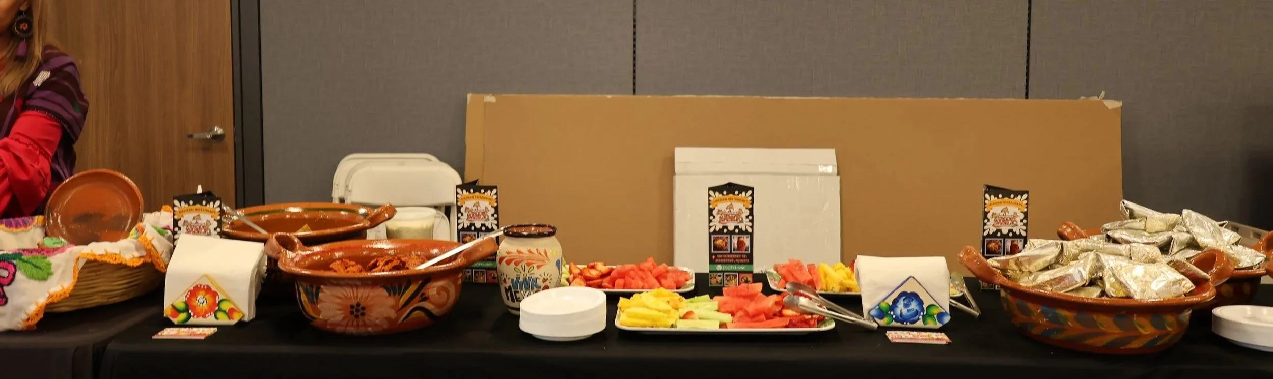 Mexican food buffet with watermelon, pineapple, and celery slices, ceramic dishes, and colorful decorations on a black table.