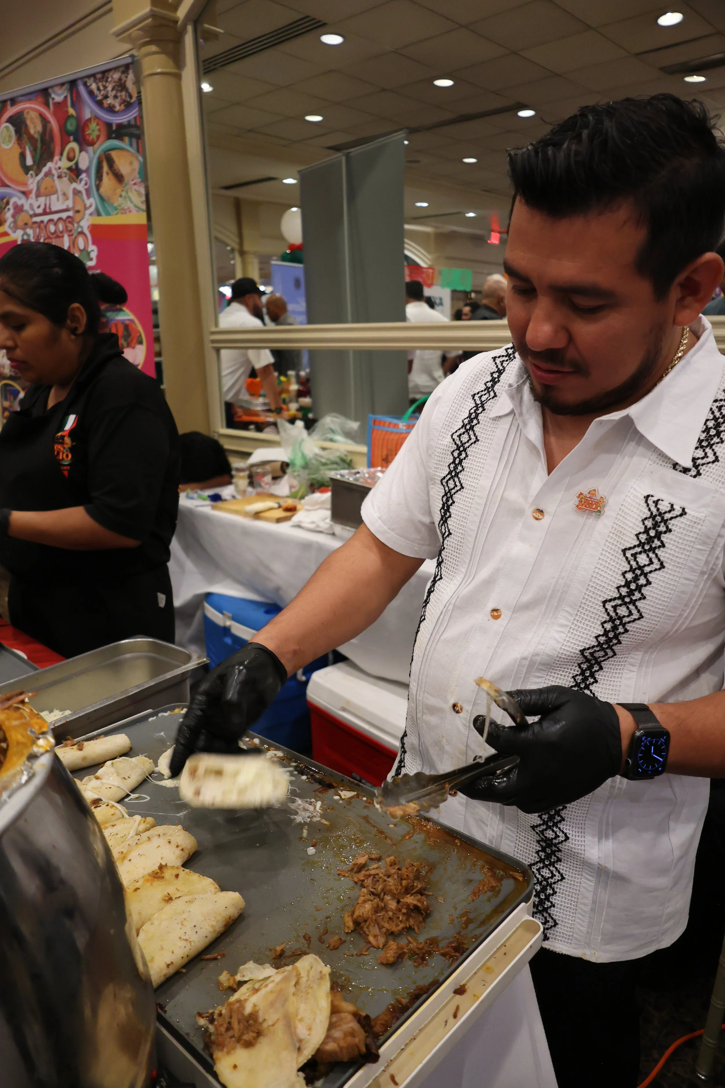 A man in a white shirt with black embroidery preparing Mexican food at a food event, with a tray of tacos, shredded meat, and tortillas. There are other people and food stalls in the background.