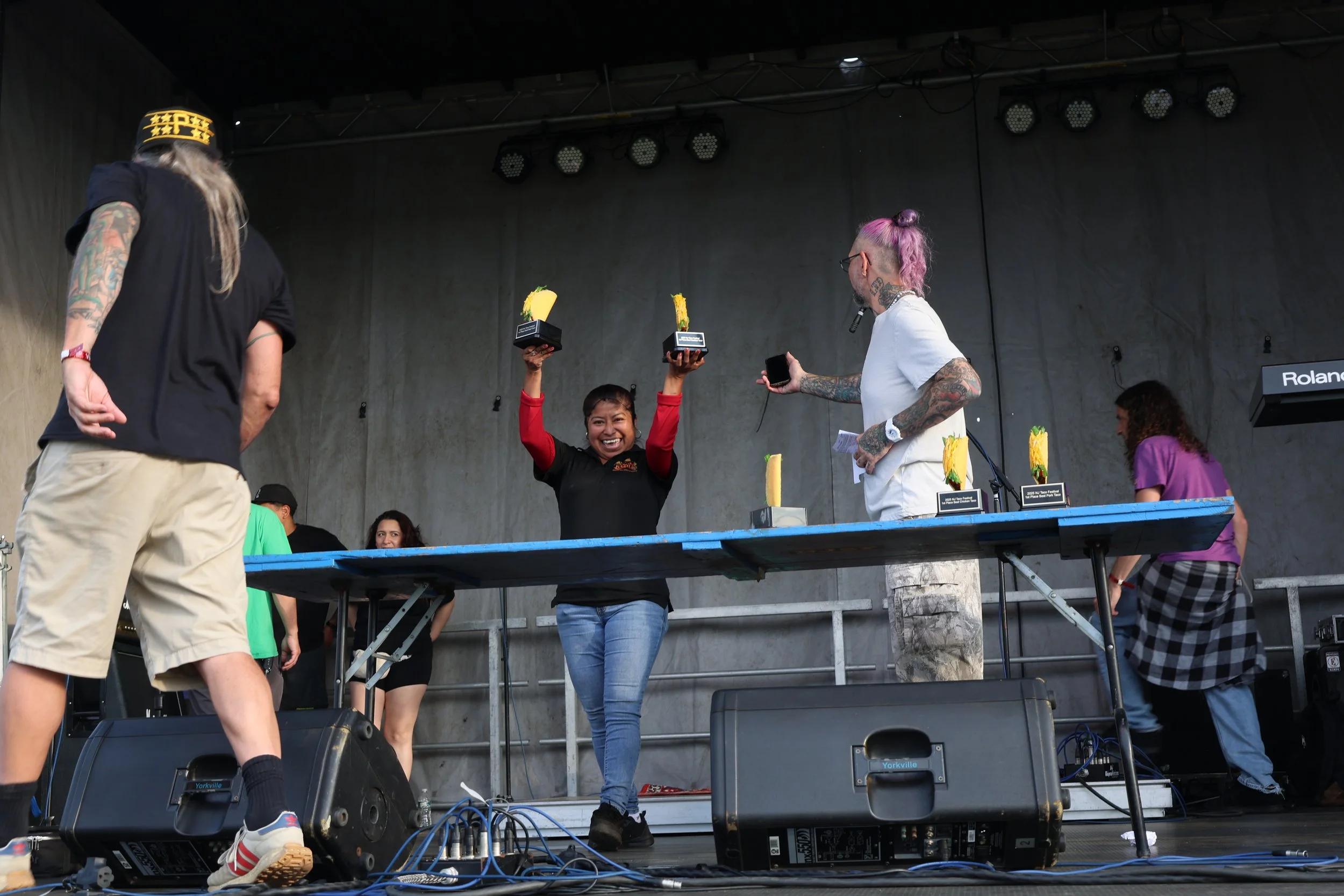 A woman with pink hair and tattoos is holding an award and smiling on stage. Another person with long hair and a cap is standing nearby. Several trophies with yellow decorations are on a table, and there is a musical keyboard in the background.