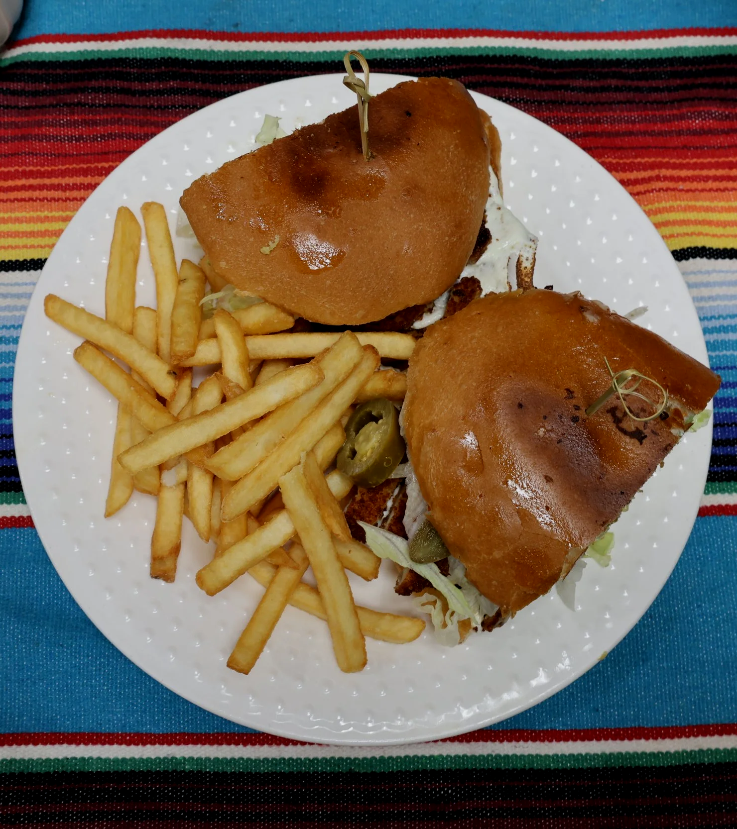 Plate of two small Mexican-style sandwiches with crispy toasted bread, filling of meat, lettuce, pickles, served with French fries and jalapeño slices on a festive colorful tablecloth.
