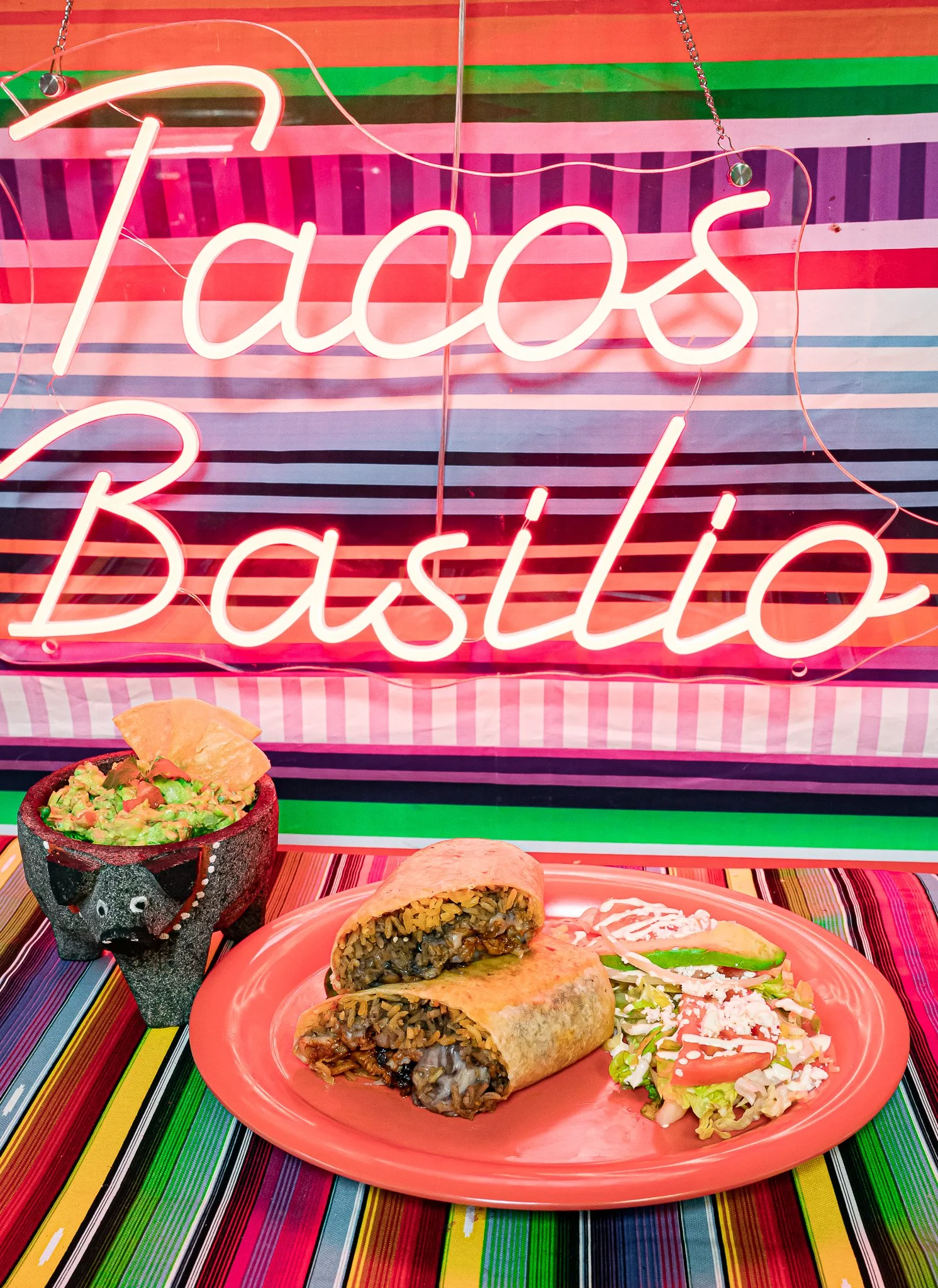 Neon sign reading 'Tacos Baja' in pink, hanging in front of striped pink, green, black, purple, white, and red backdrop. A plate with two rolled tacos filled with cooked mushrooms and onions, a side salad with lettuce, tomato, and cheese, along with 