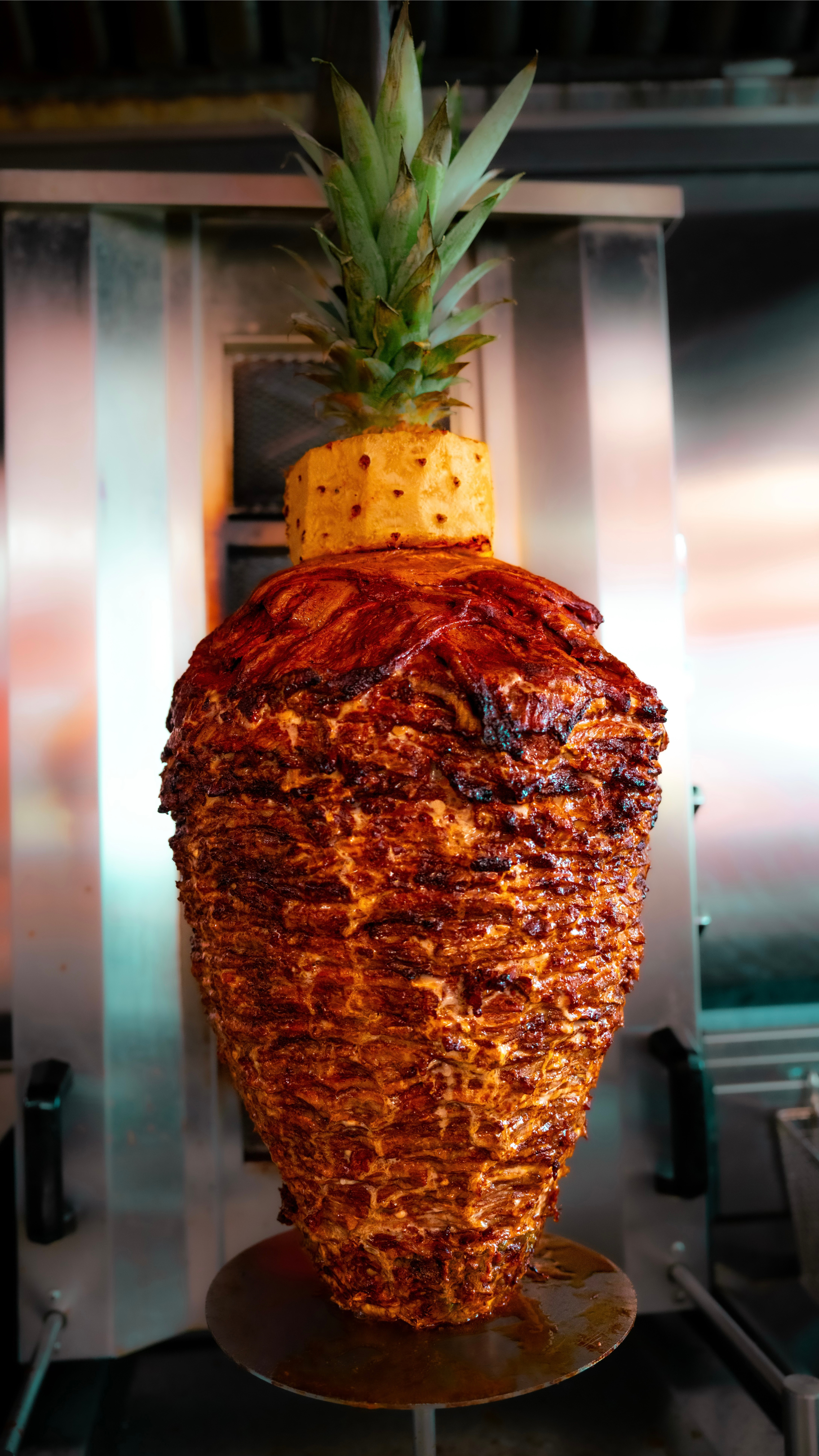 A freshly cooked whole pineapple wrapped in layers of seasoned meat, skewered on a vertical rotisserie, ready for slicing.