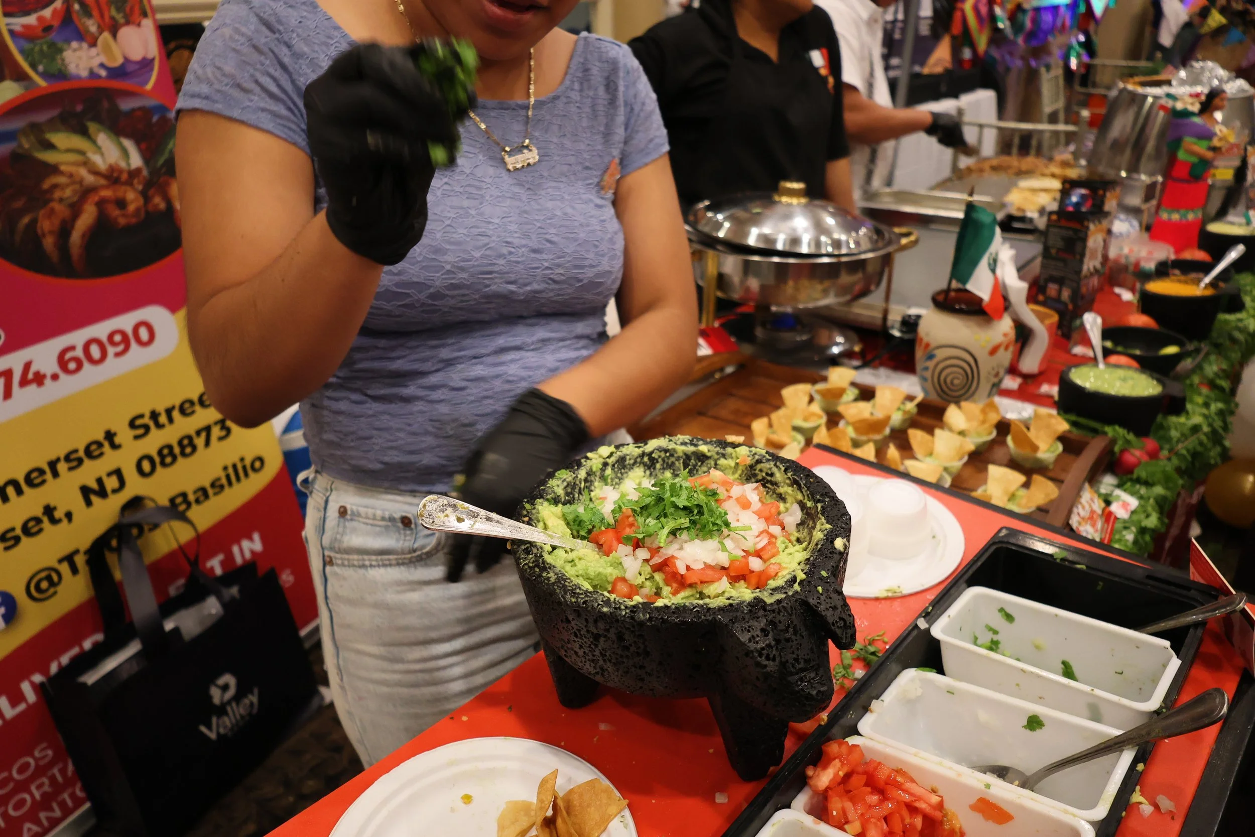 A woman preparing Mexican food at a market stall, with ingredients like chopped tomatoes, avocado, and tortilla chips displayed on the table.