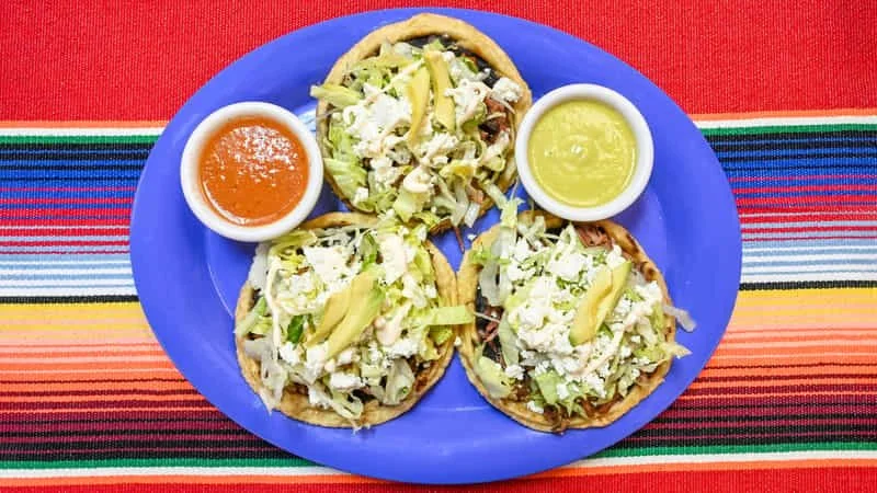 Three tacos topped with shredded lettuce and cheese on a blue plate, with two small bowls of salsa and guacamole on a colorful striped tablecloth.