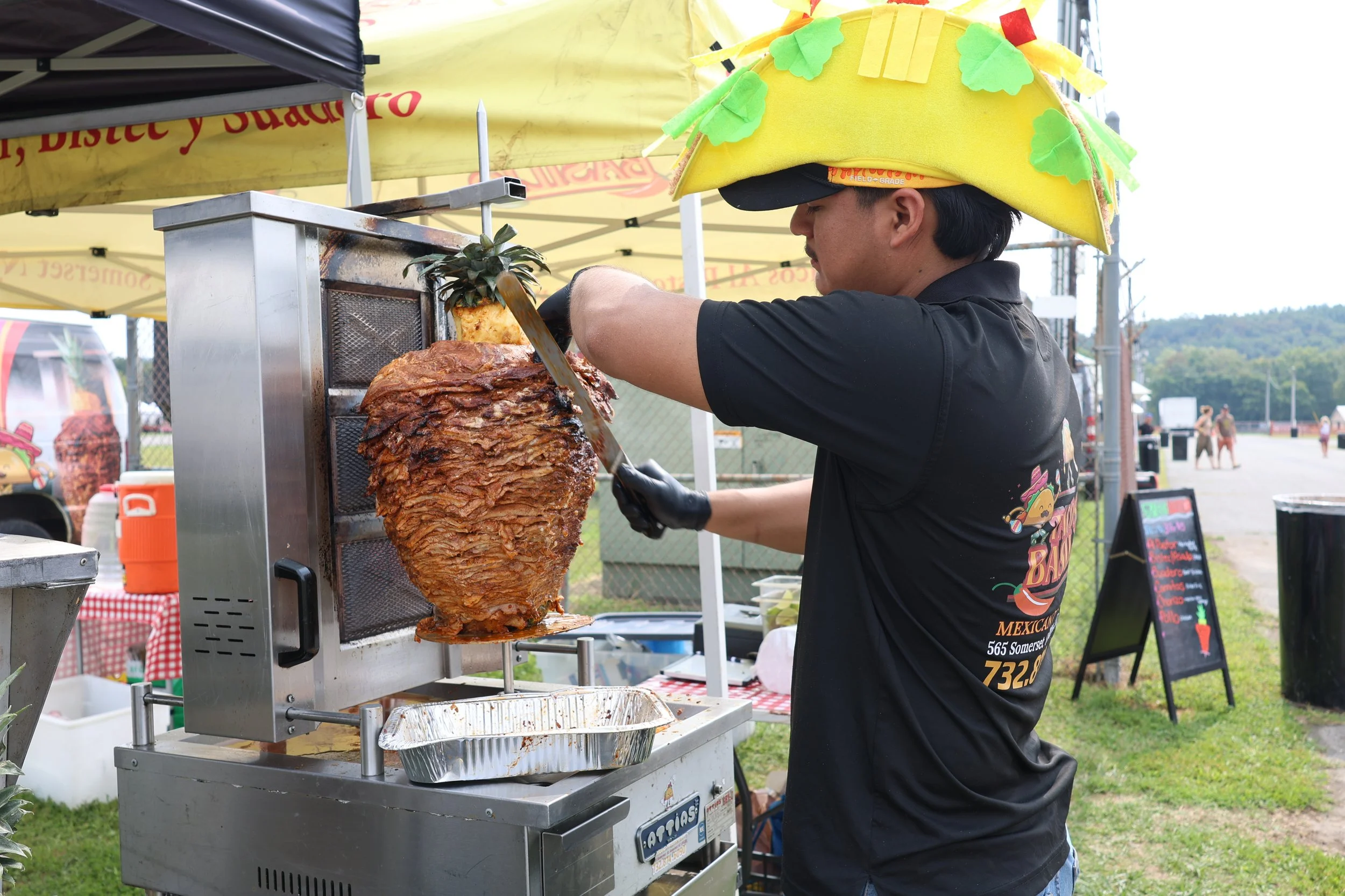 A man wearing a black shirt and a yellow hat with green leaves is grilling a large stack of sliced meat, possibly pork, on a vertical rotisserie at an outdoor food stand.