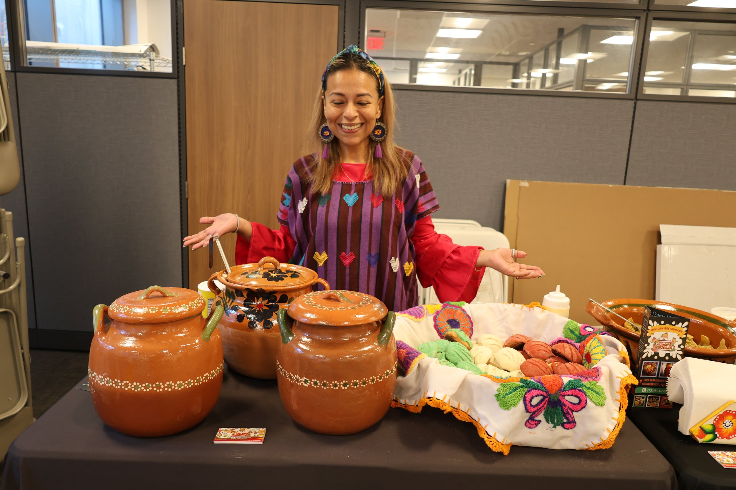 A woman with light brown hair, wearing a colorful headscarf, large embroidered earrings, and a vibrant embroidered shirt, stands behind a table with traditional Mexican pottery and colorful textiles, smiling with her hands open in a cheerful gesture.